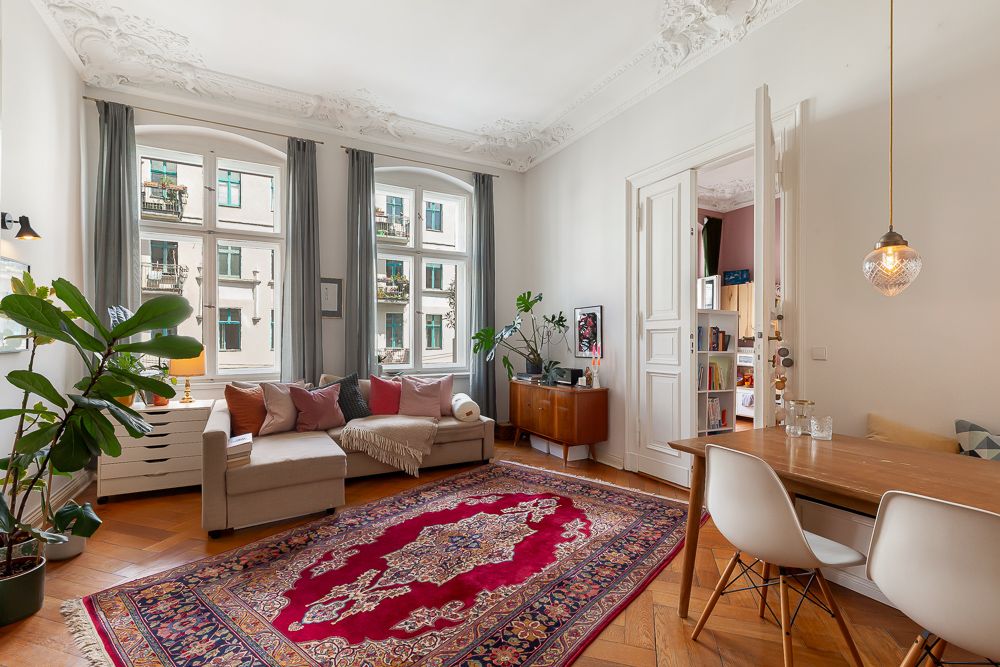 Helles Wohnzimmer mit Sofa, Teppich, Pflanzen, Esstisch und großen Fenstern.
