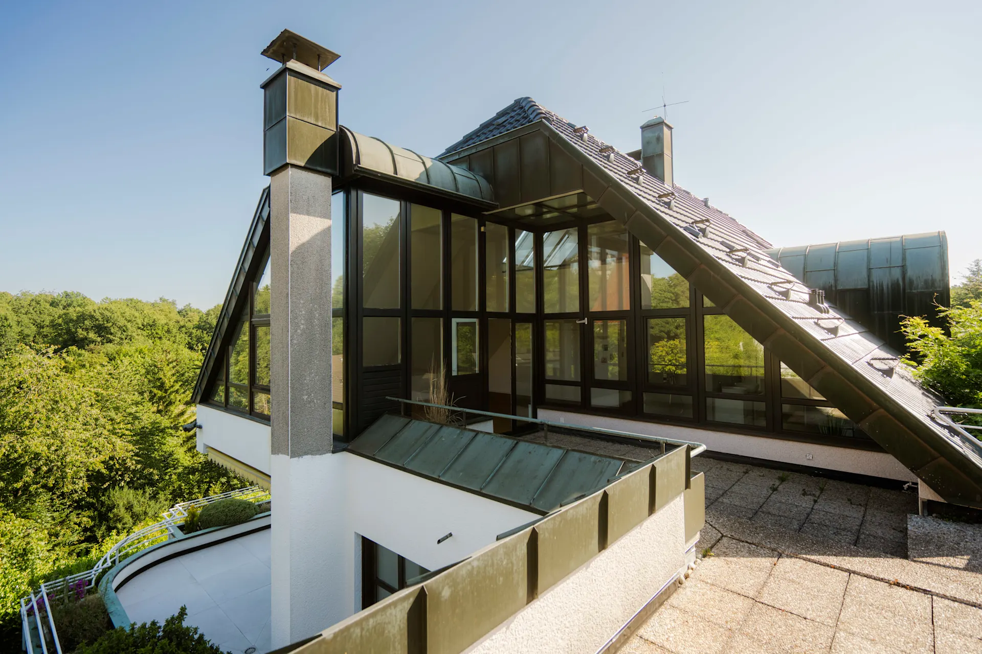Moderne Wohnhaus-Fassade mit großen Fenstern und schrägen Dächern, umgeben von Bäumen.