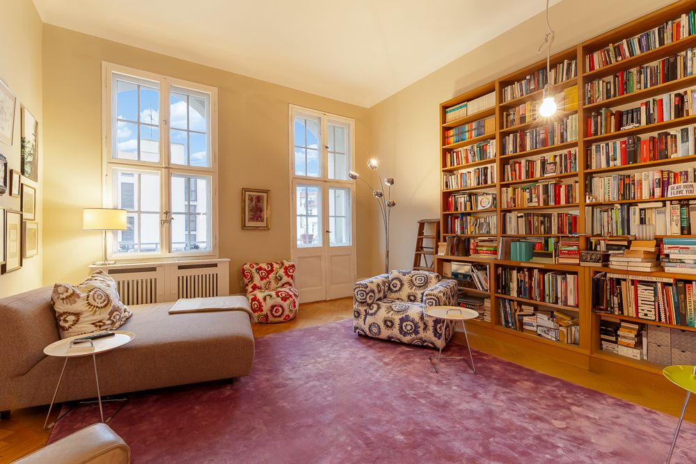 Helles Wohnzimmer mit großen Fenstern, bunten Sesseln, Bücherwand und lila Teppich.