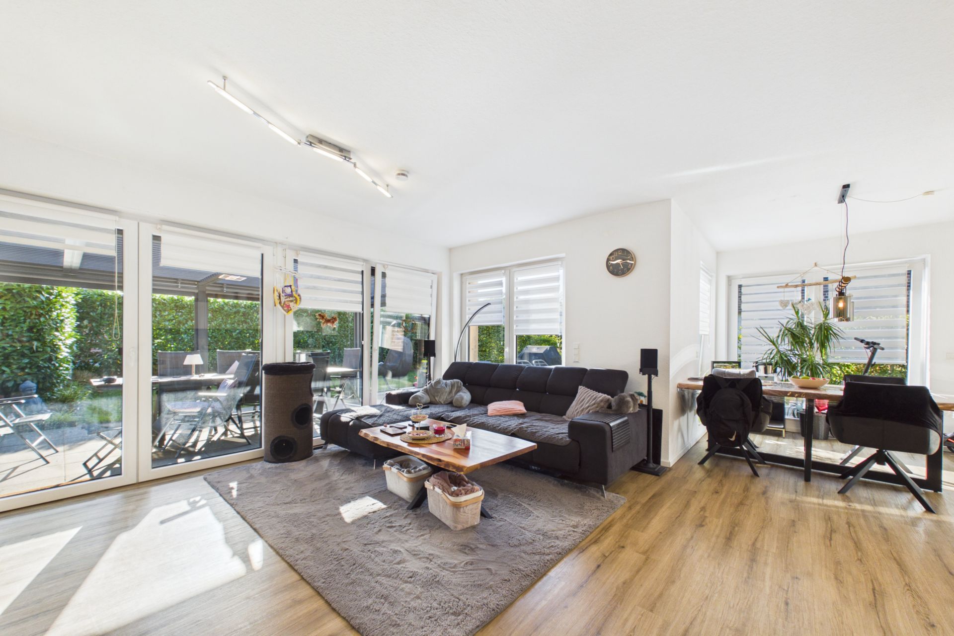 Helles Wohnzimmer mit Sofa, Couchtisch, Teppich und angrenzendem Essbereich vor großen Fenstern.