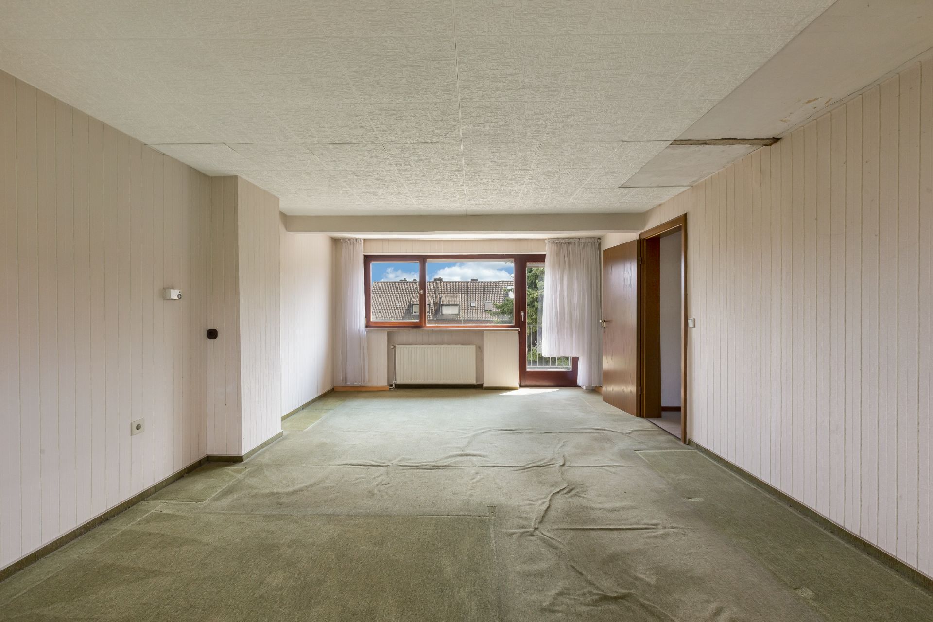 Leeres Zimmer mit grüner Auslegeware, großen Fenstern und Zugang zum Balkon.