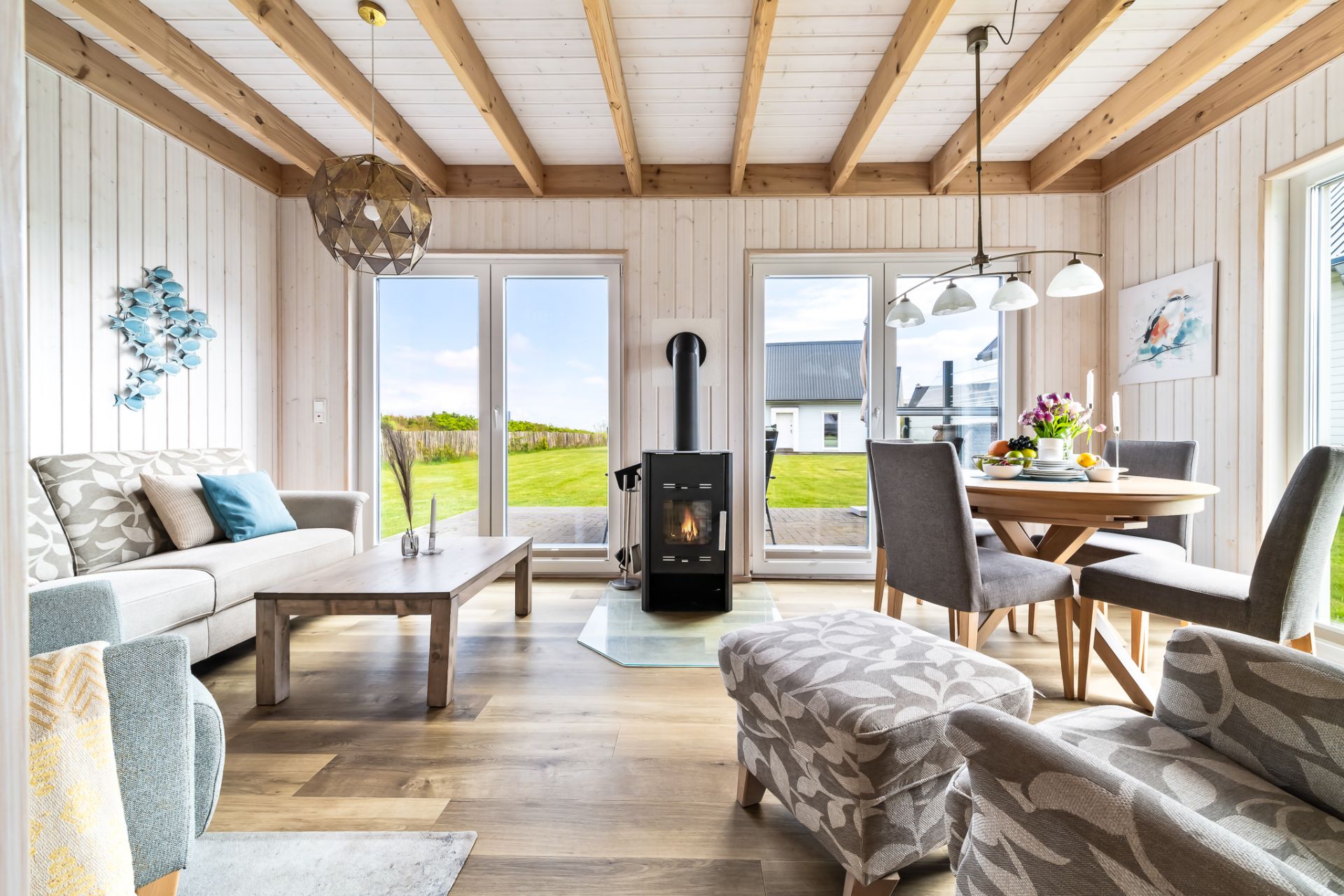 Helles Wohnzimmer mit Sofa, Sesseln, Holzdecke, Kaminofen und Esstisch vor großen Fenstern mit Gartenblick.