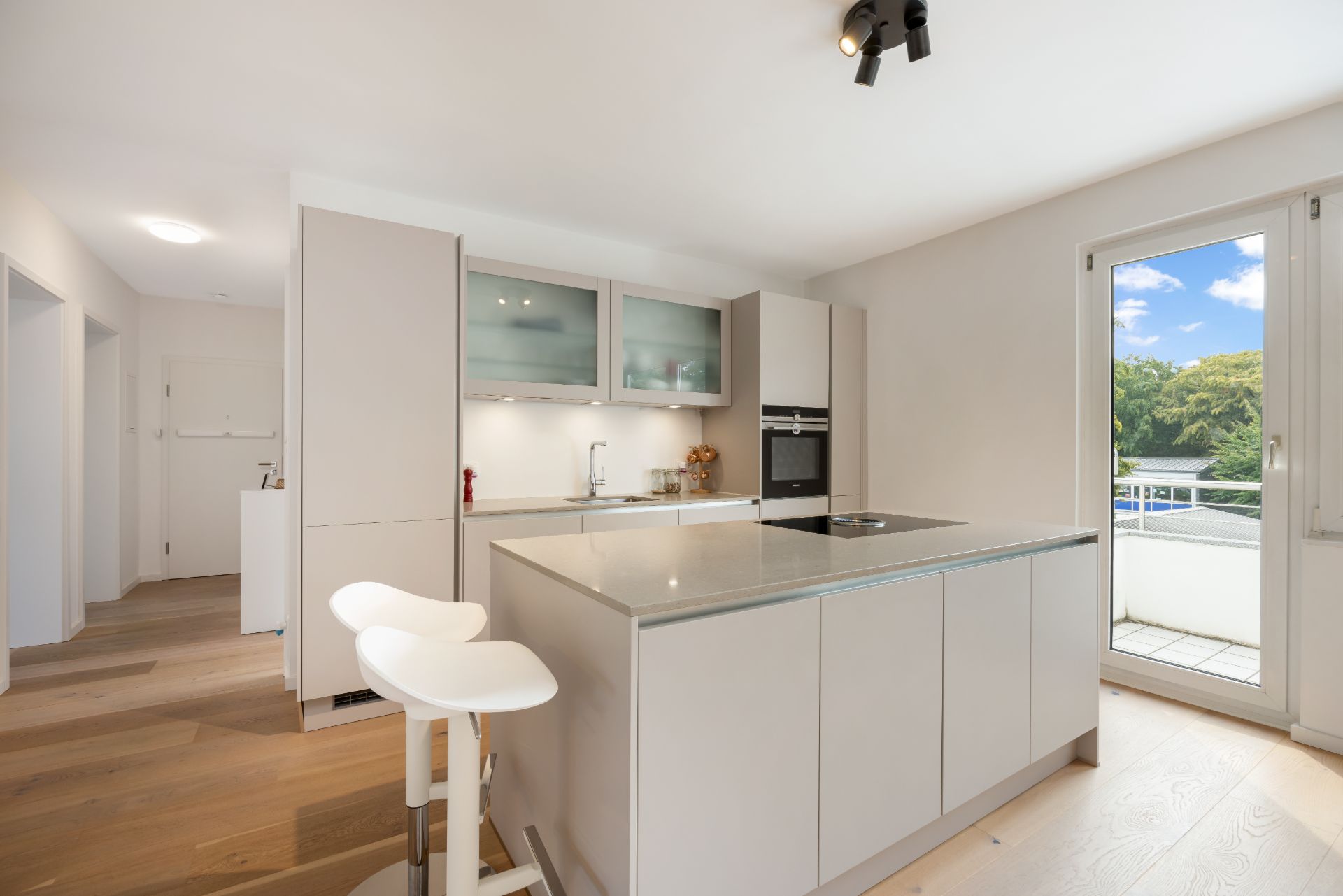Helle, moderne Küche mit Kochinsel, zwei weißen Barhockern und Zugang zum Balkon.
