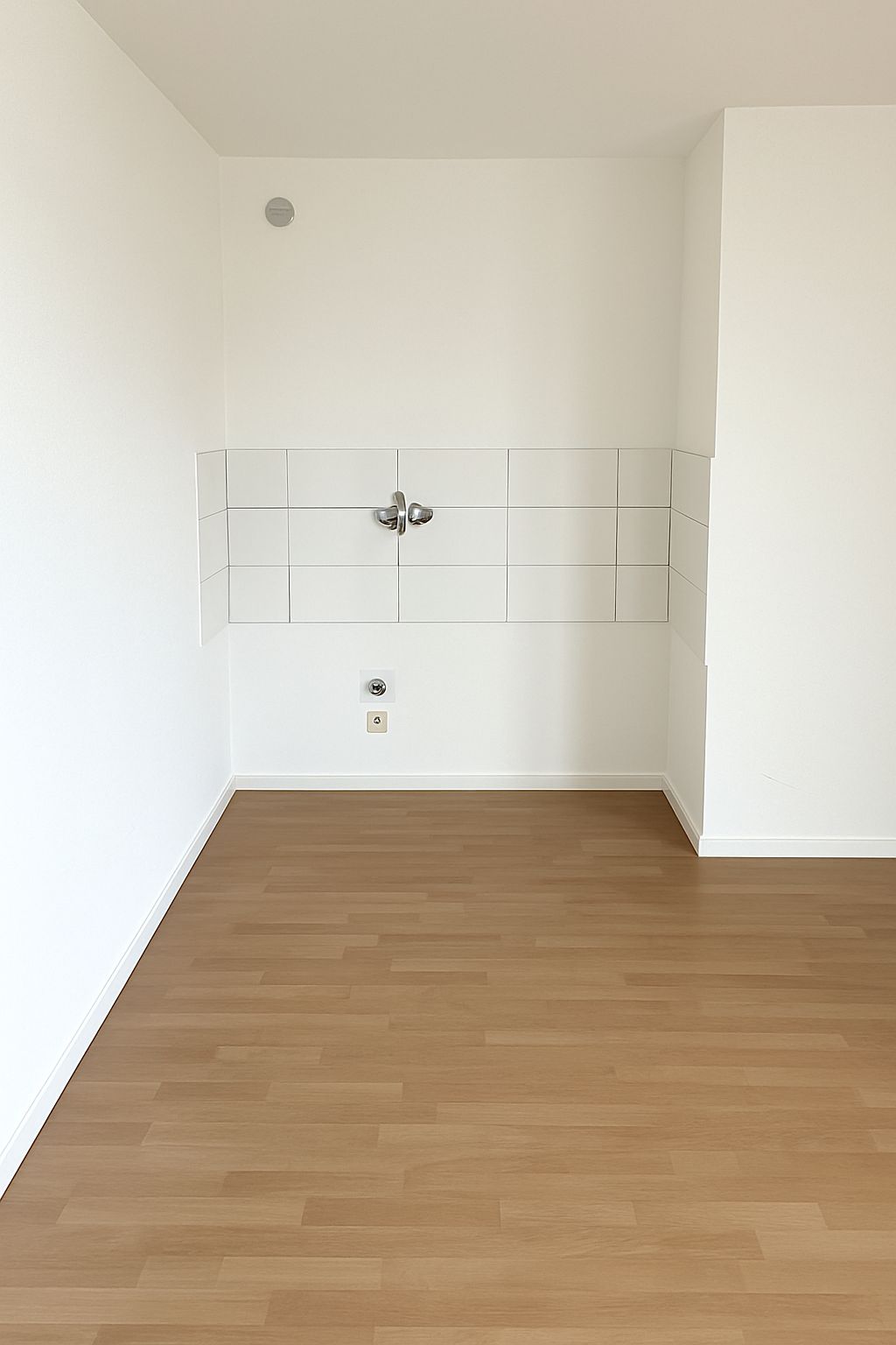 Leerer Raum mit hellem Laminatboden und weißen Fliesen an der Wand mit Wasseranschlüssen.