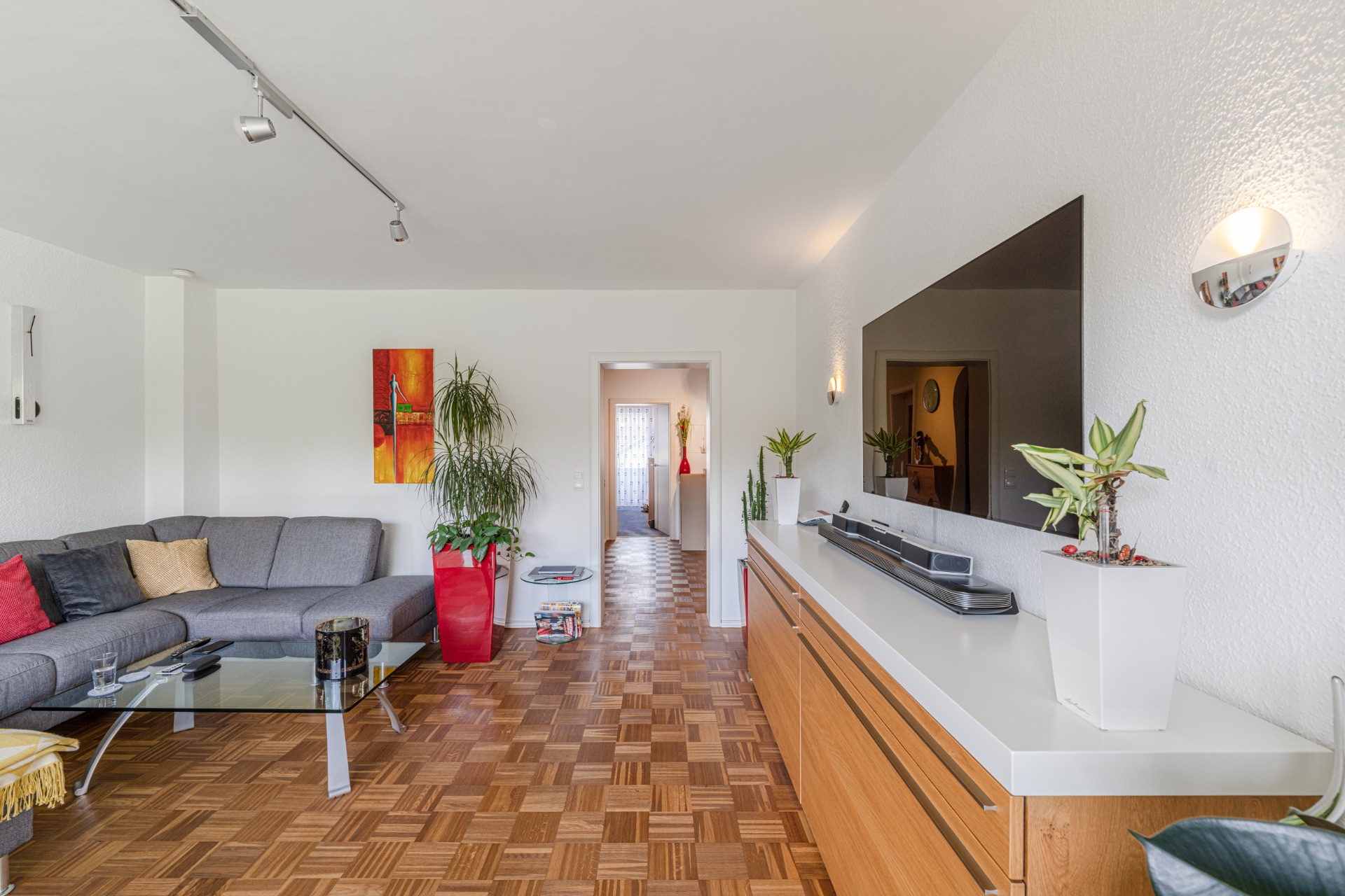 Modernes Wohnzimmer mit grauem Ecksofa, Glastisch, großen Pflanzen und Fernseher an der Wand.