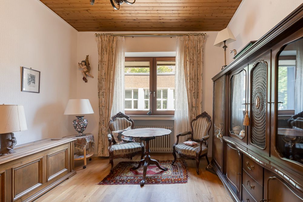 Wohnzimmer mit Holzmöbeln, zwei Stühlen, rundem Tisch, Teppich und Fenster mit Vorhängen.