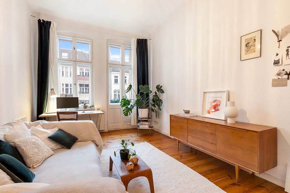 Helles Wohnzimmer mit Sofa, Holztisch, Sideboard, Schreibtisch und großen Fenstern.