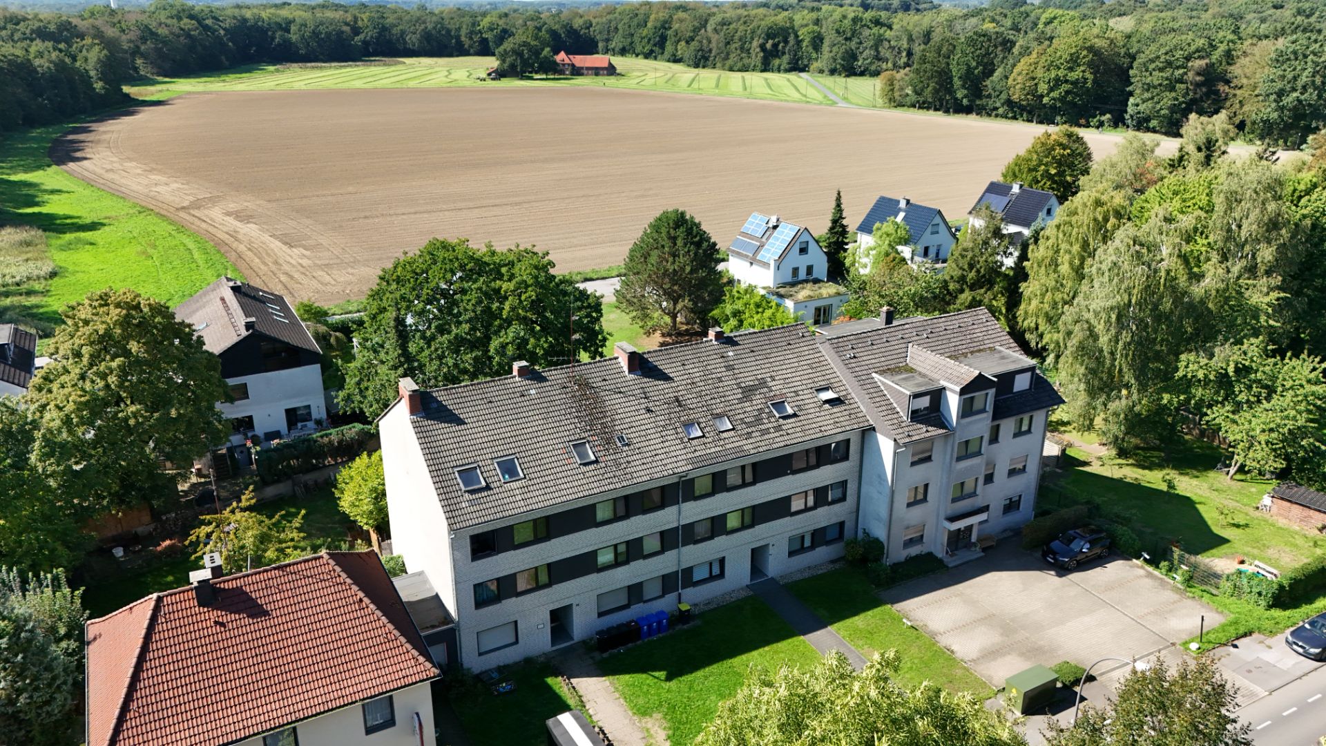 Mehrfamilienhaus mit grauem Dach, Grünfläche und Parkplätzen, im Hintergrund ein Feld und Bäume.