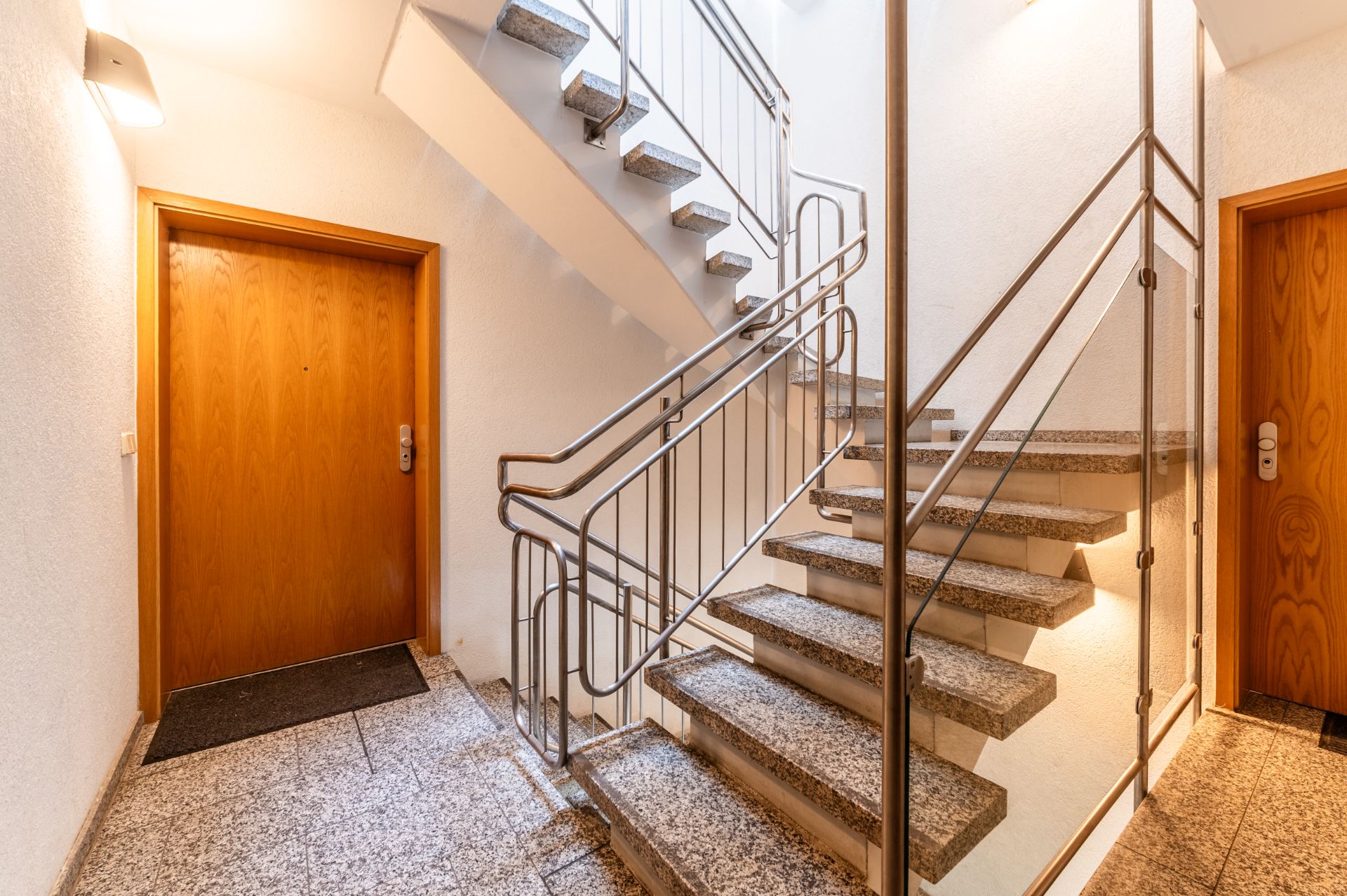 Helles Treppenhaus mit Granitstufen, Metallgeländer und zwei Holztüren.