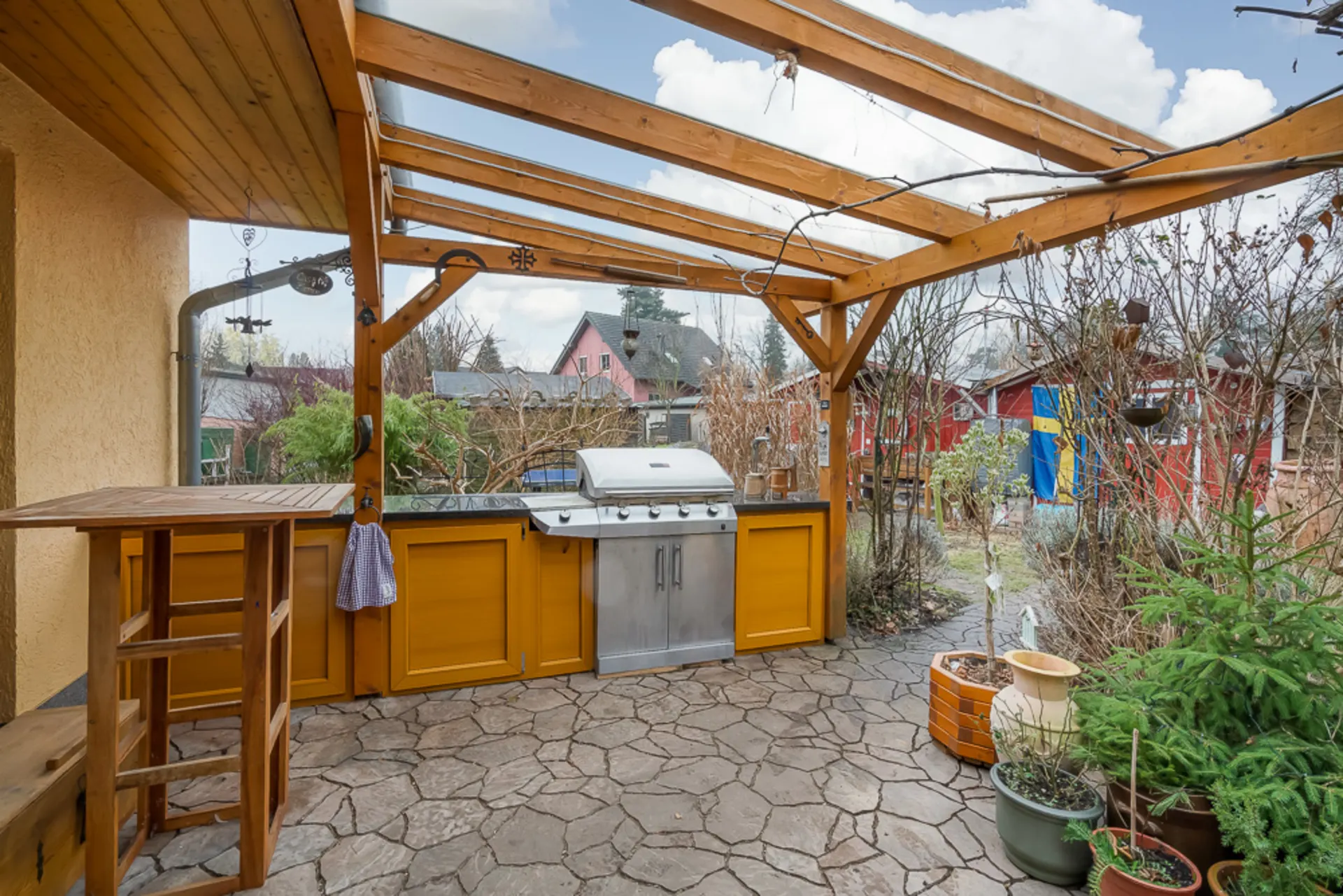 Überdachte Terrasse mit Grill, gelben Schränken, Sitzmöbeln und Pflanzen im Garten.