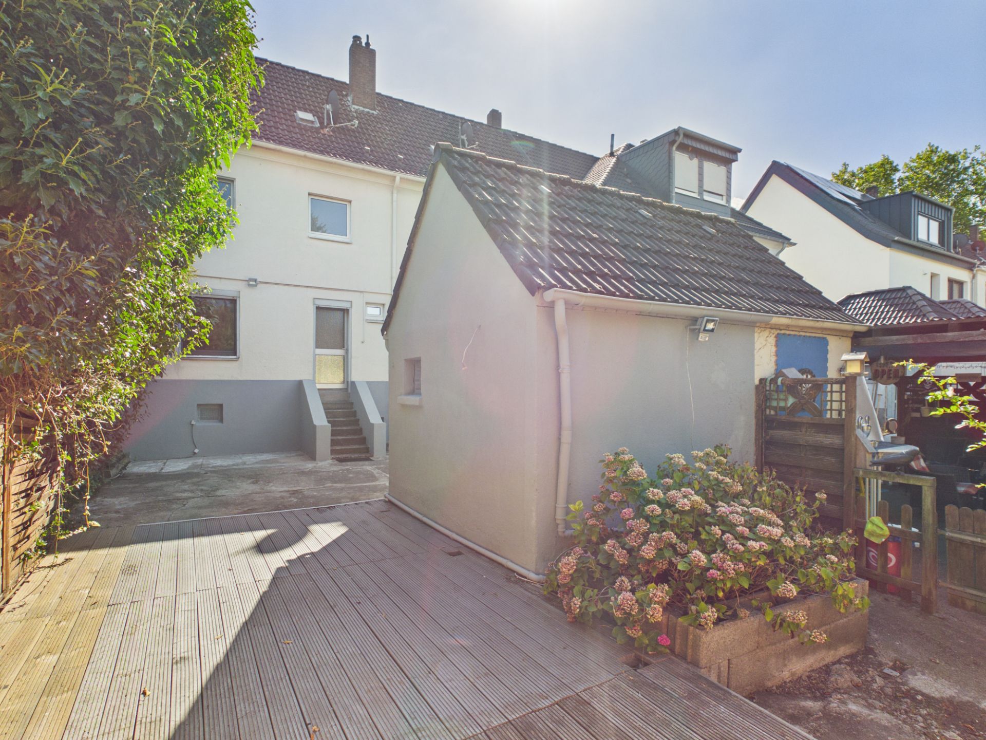 Gepflasterter Hinterhof mit kleinem Anbau, Holzterrasse und Blumenbeet vor Wohnhaus.
