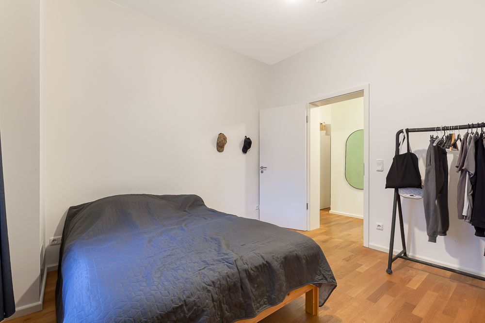 Schlafzimmer mit Bett, Kleiderständer und offener Tür, Holzboden und weiß gestrichene Wände.