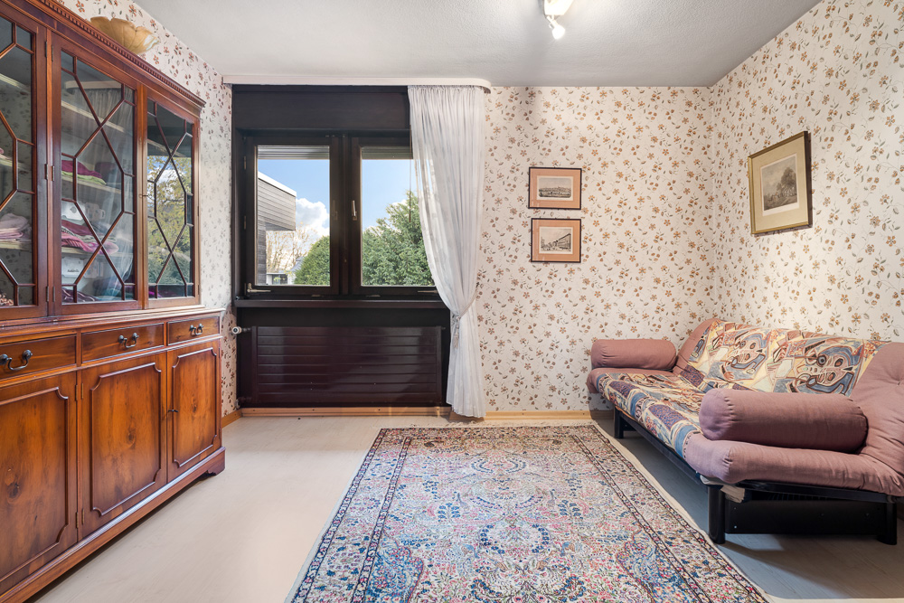Wohnzimmer mit floral tapezierter Wand, Sofa, Holzschrank, Teppich und Fenster mit Blick ins Grüne.