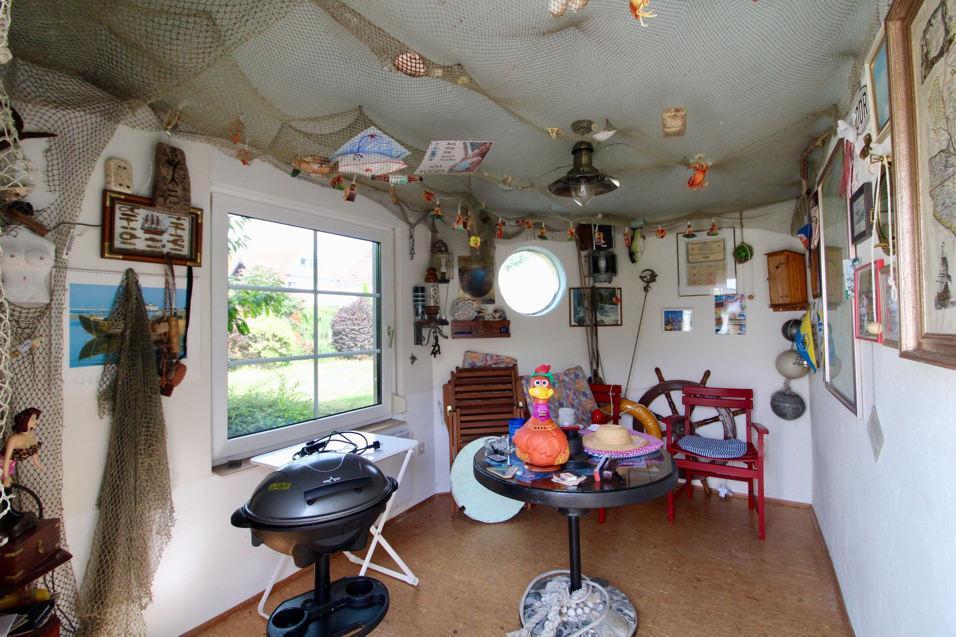 Kleiner Raum mit maritimer Dekoration, rundem Fenster, Tisch, Grill und roten Stühlen.
