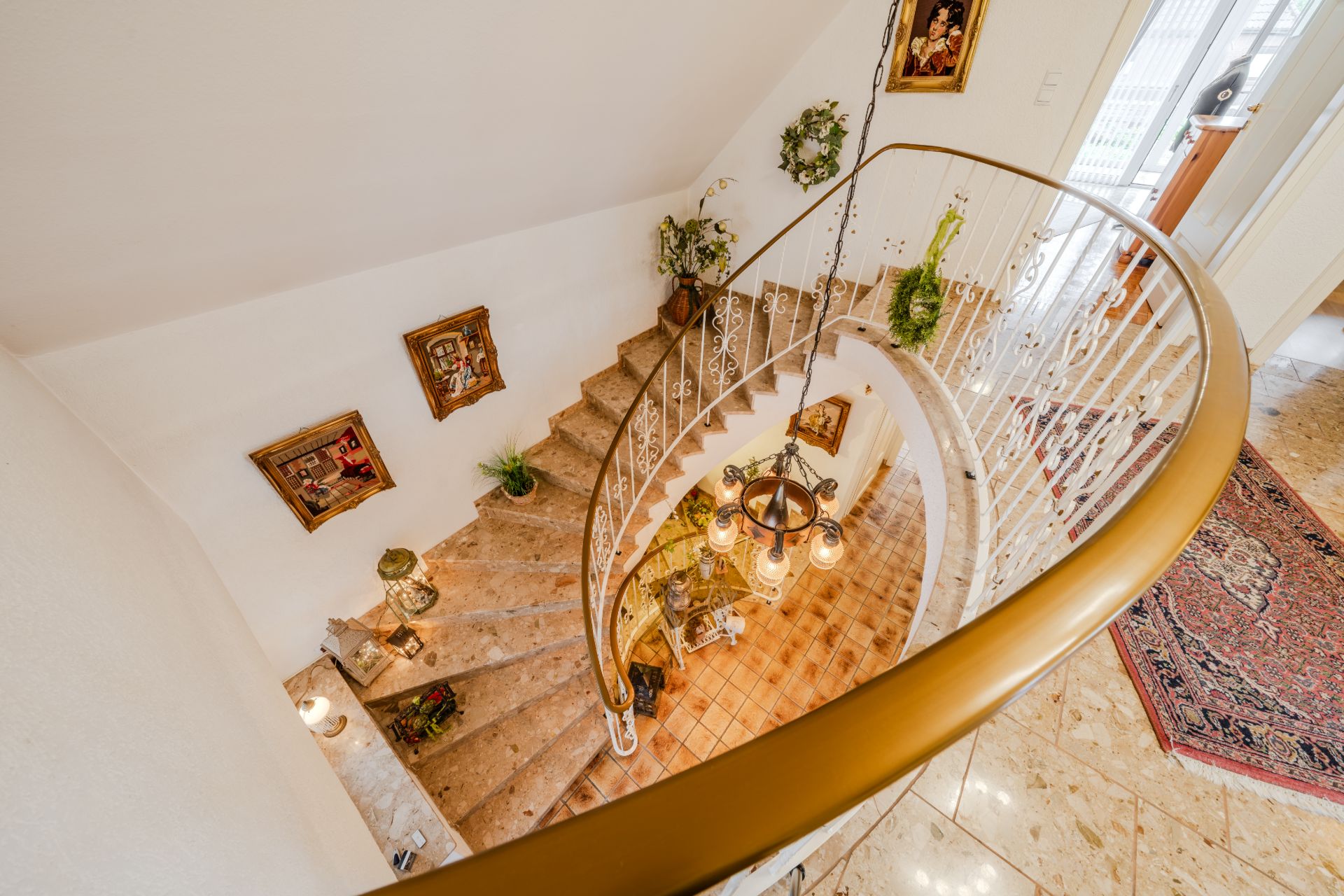 Blick von oben auf eine geschwungene Treppe mit Dekorationen und Gemälden an den Wänden.