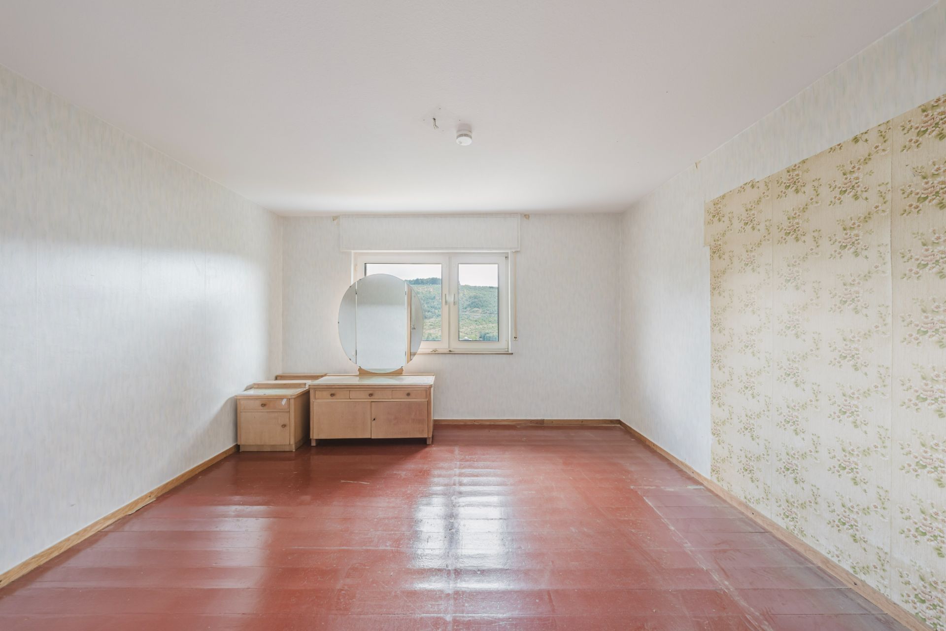 Leeres Zimmer mit rotbraunem Boden, zwei Holzmöbeln und Fenster mit Ausblick.