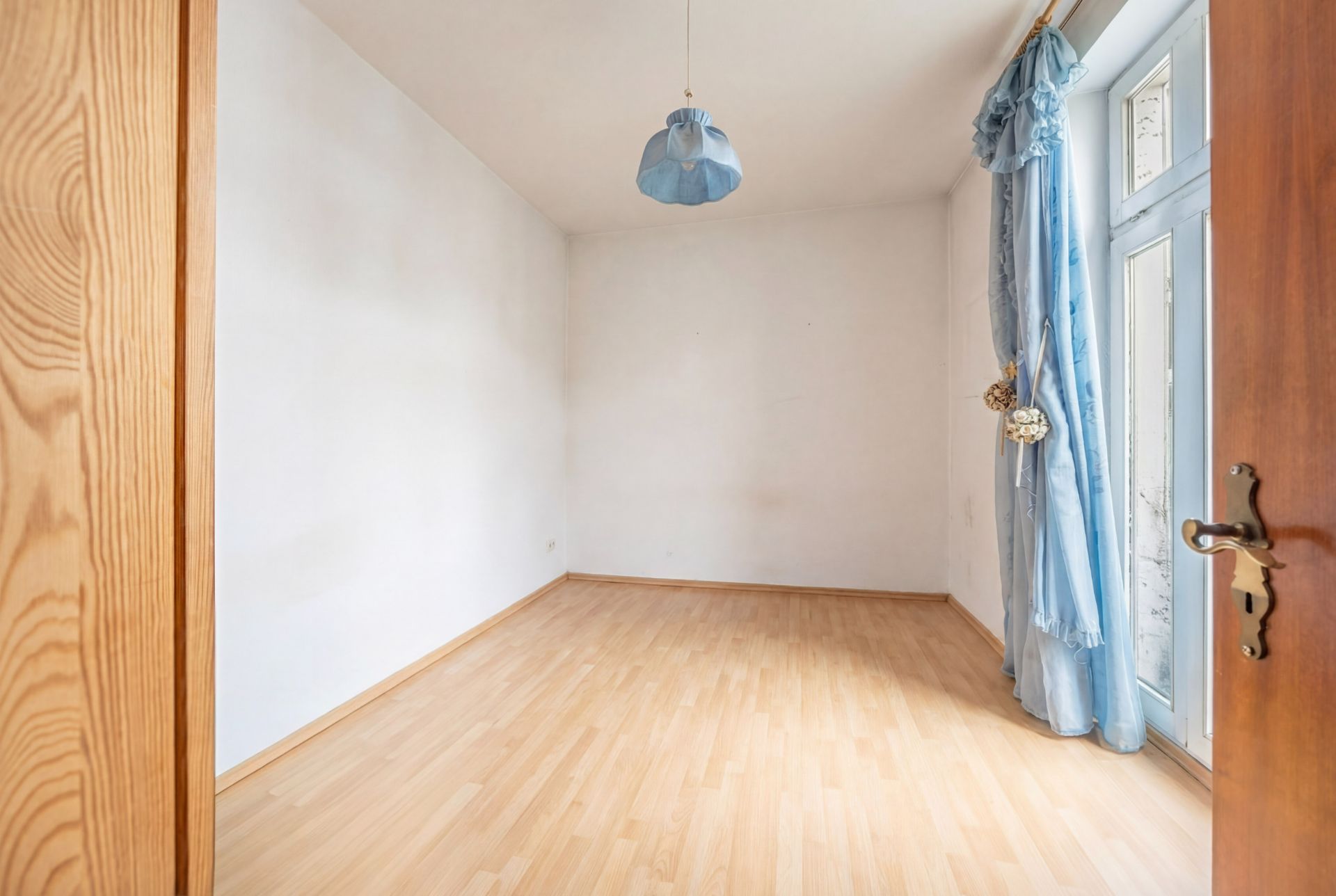 Leeres Zimmer mit hellem Holzboden, weißer Wand, blauer Lampe und blauen Vorhängen an einer Glastür.