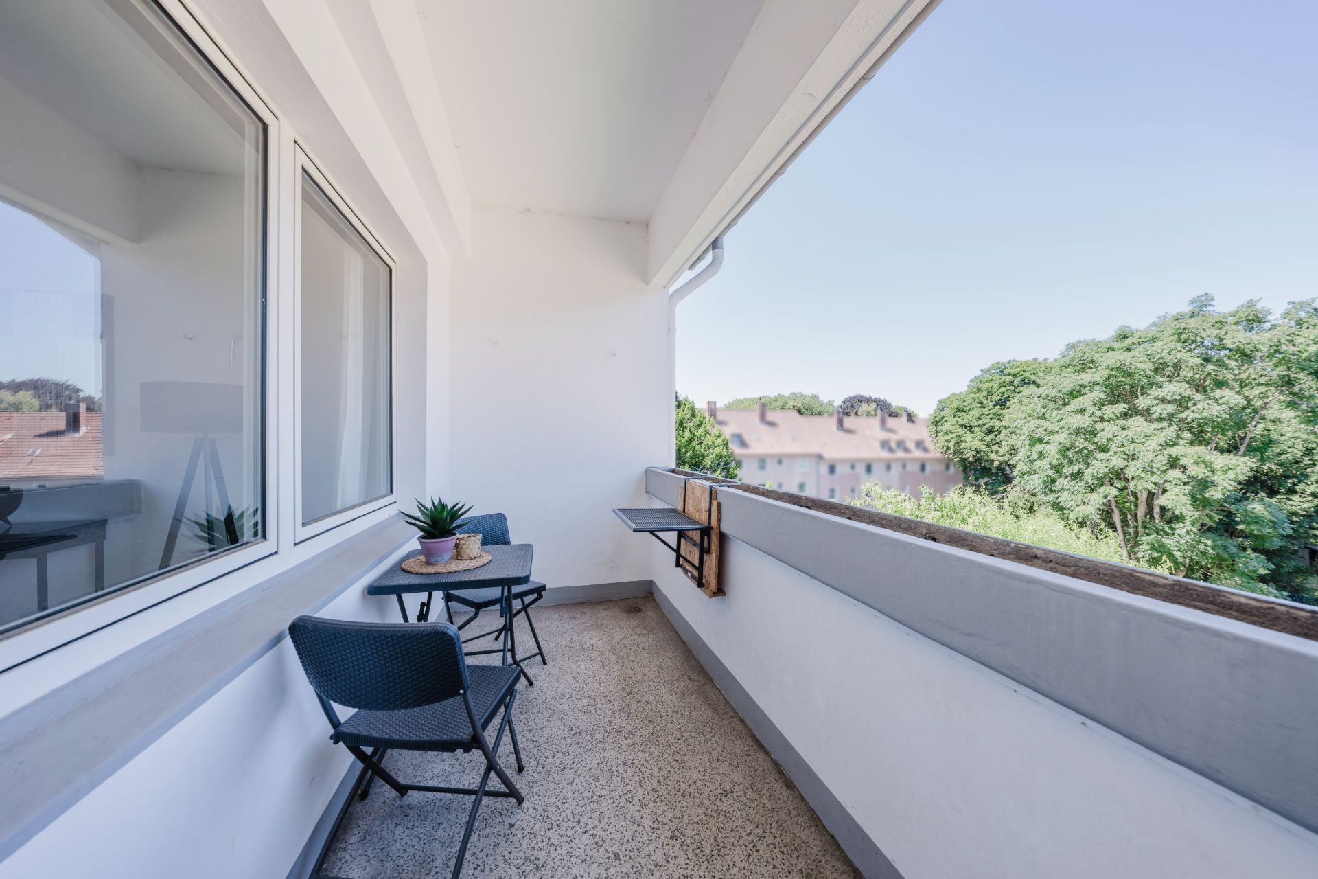 Balkon mit zwei Stühlen, kleinem Tisch und Blick auf Bäume sowie Nachbargebäude.