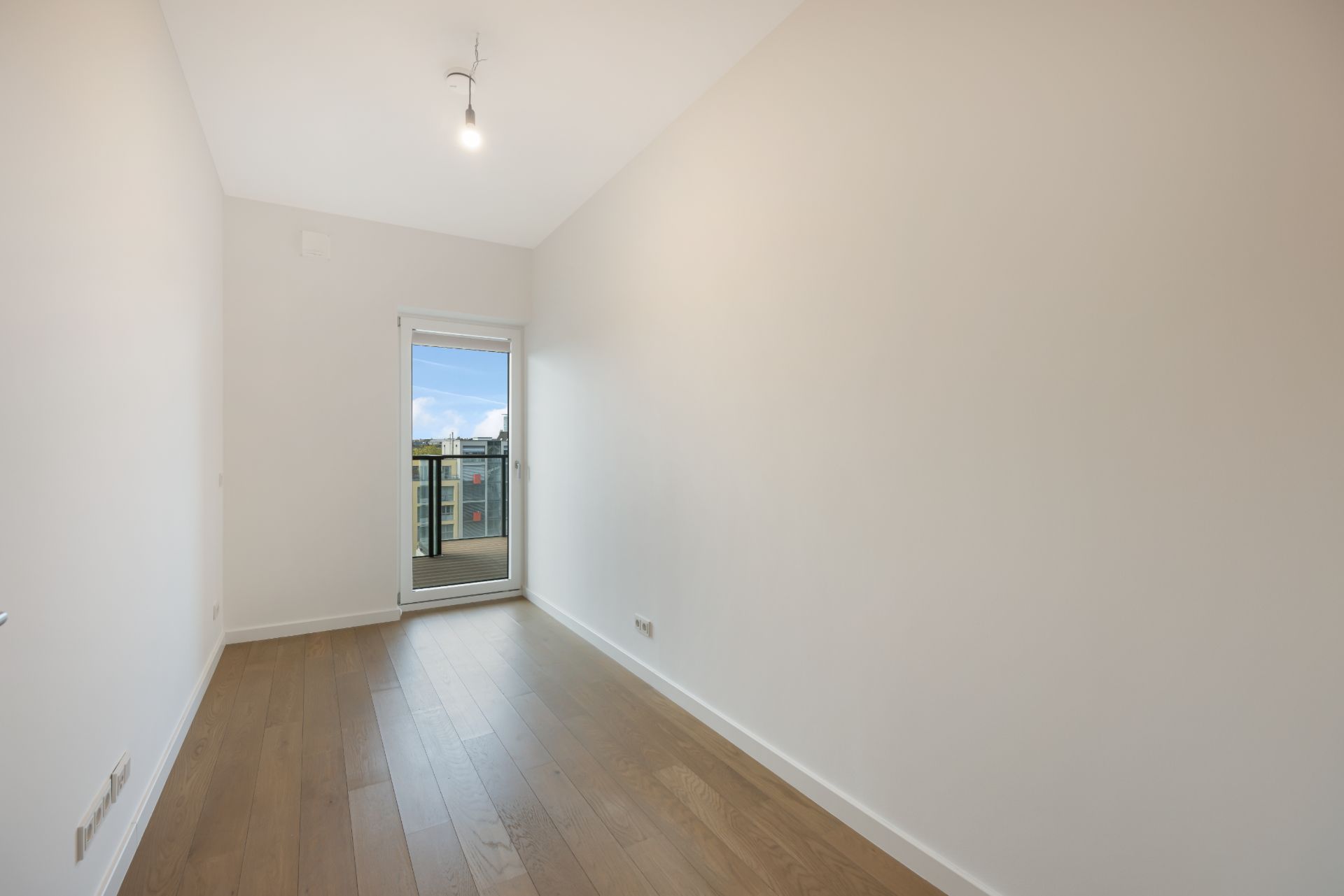 Leerer, schmaler Raum mit Holzboden und Glastür zu einem Balkon.