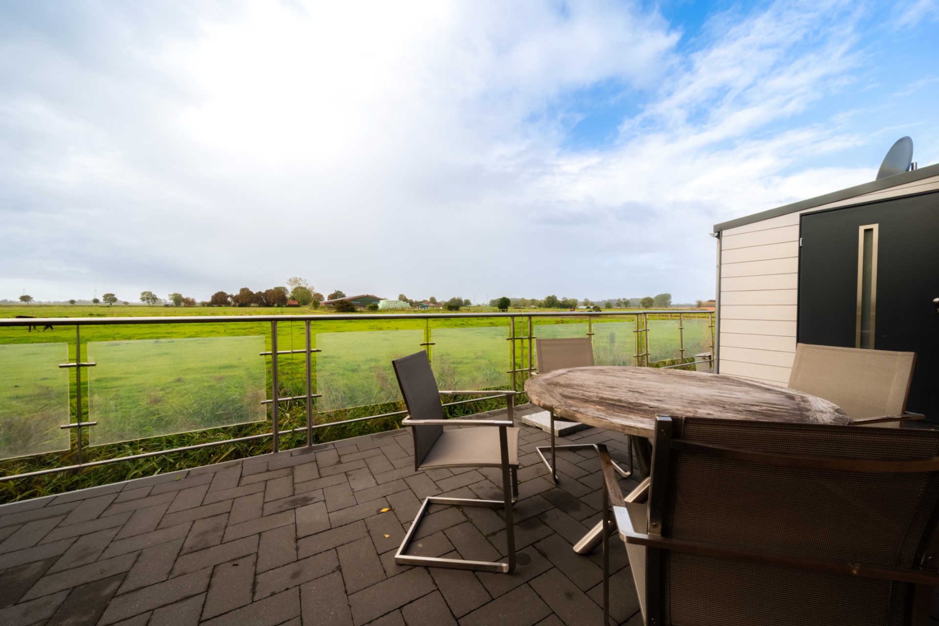 Terrasse mit rundem Holztisch und vier Stühlen, Blick auf grüne Wiese und Himmel.