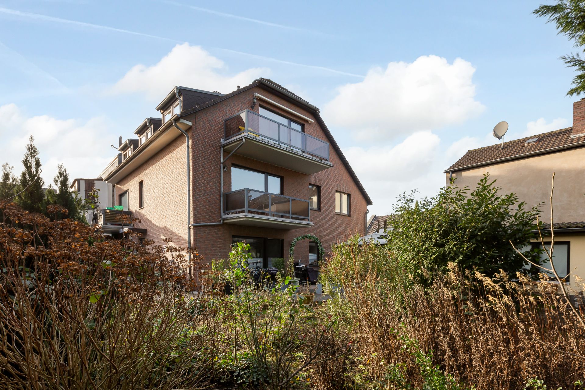 Mehrfamilienhaus mit Balkonen, umgeben von Gartenpflanzen, unter blauem Himmel.