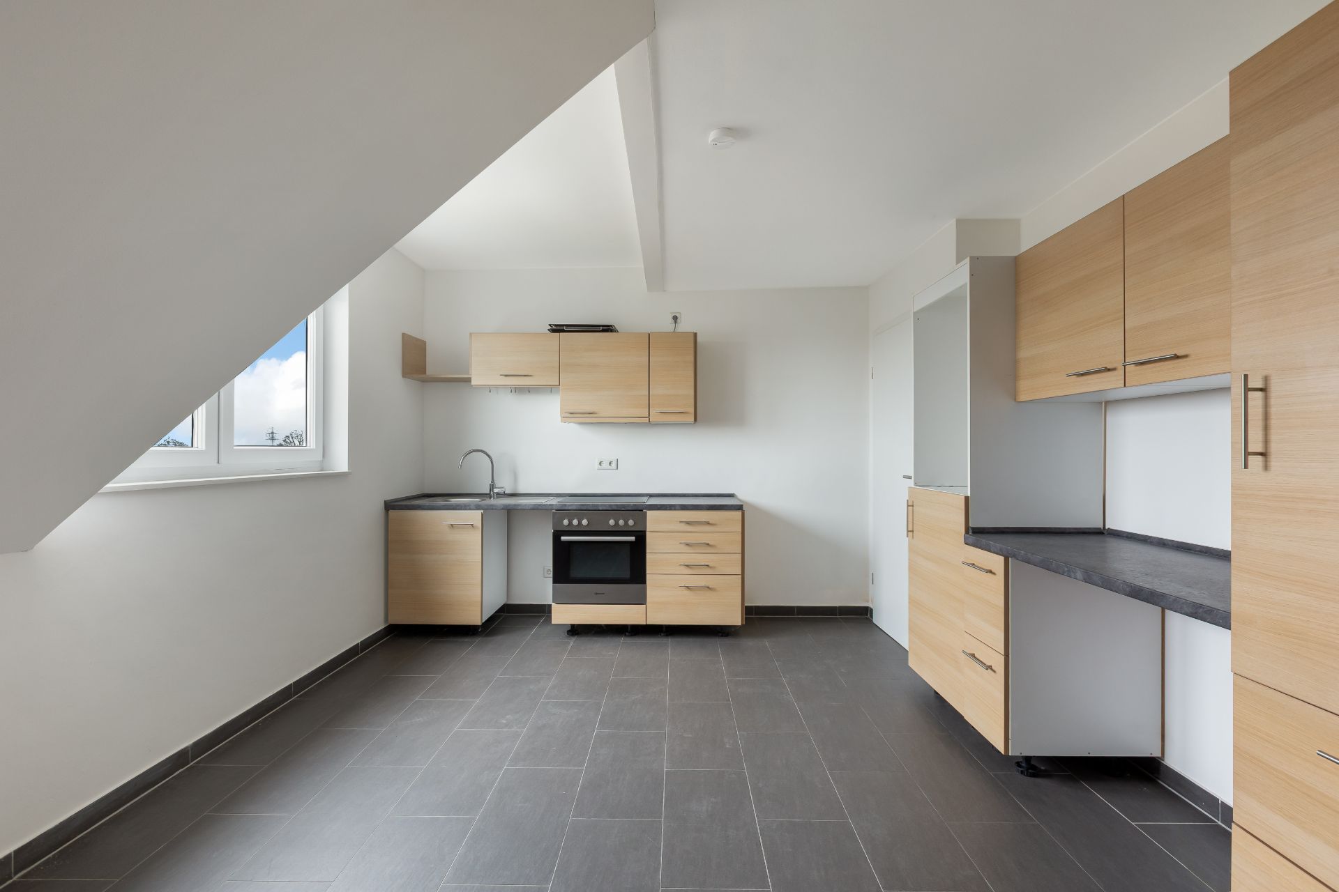 Moderne, leere Küche mit hellen Holzmöbeln und grauem Fliesenboden unter Dachschräge.