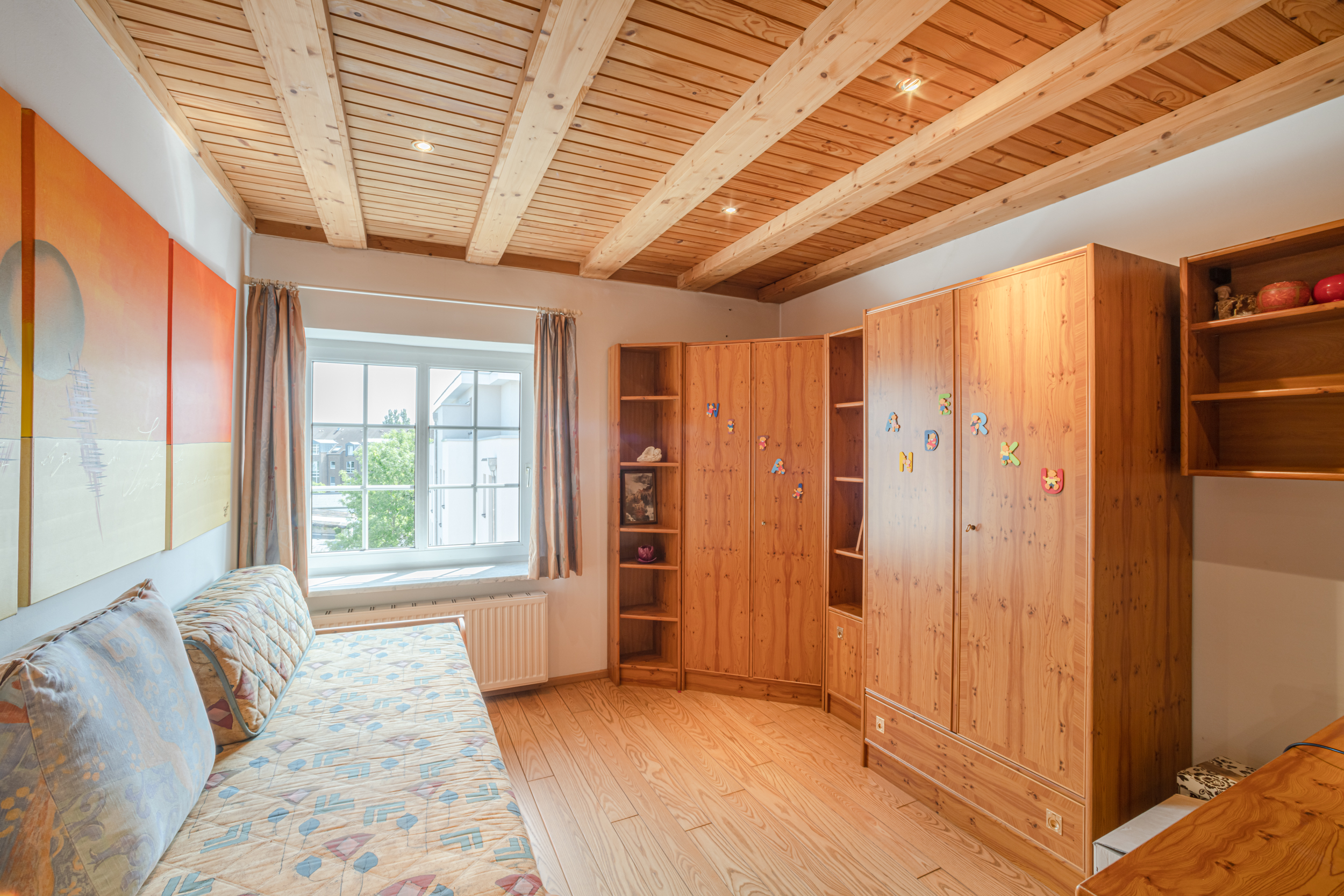 Helles Zimmer mit Holzbalkendecke, Bett, großen Holzschränken und Fenster mit Vorhängen.