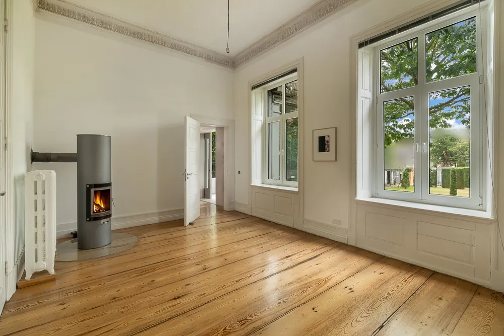 Helles Zimmer mit Holzboden, Kaminofen, zwei großen Fenstern und offener Tür.