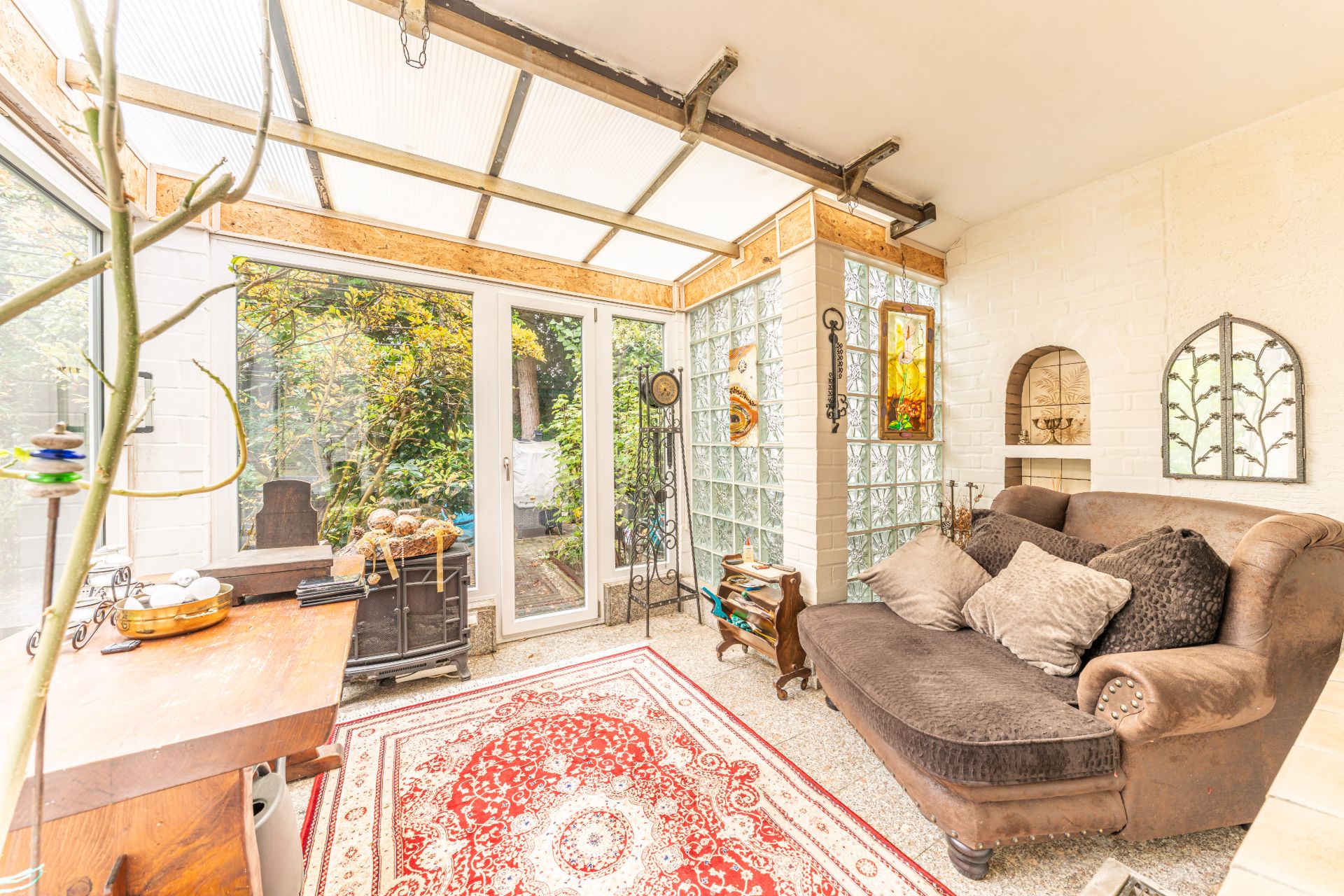 Heller Wintergarten mit Sofa, rotem Teppich und Blick in den Garten durch große Glastüren.
