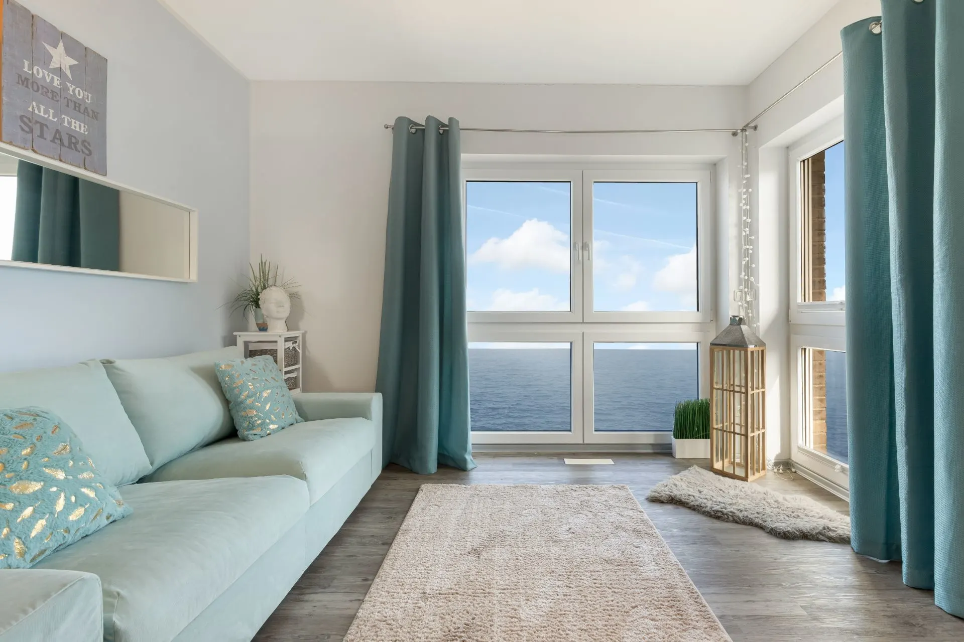 Helles Wohnzimmer mit mintfarbenem Sofa, Teppich und großen Fenstern mit Meerblick.