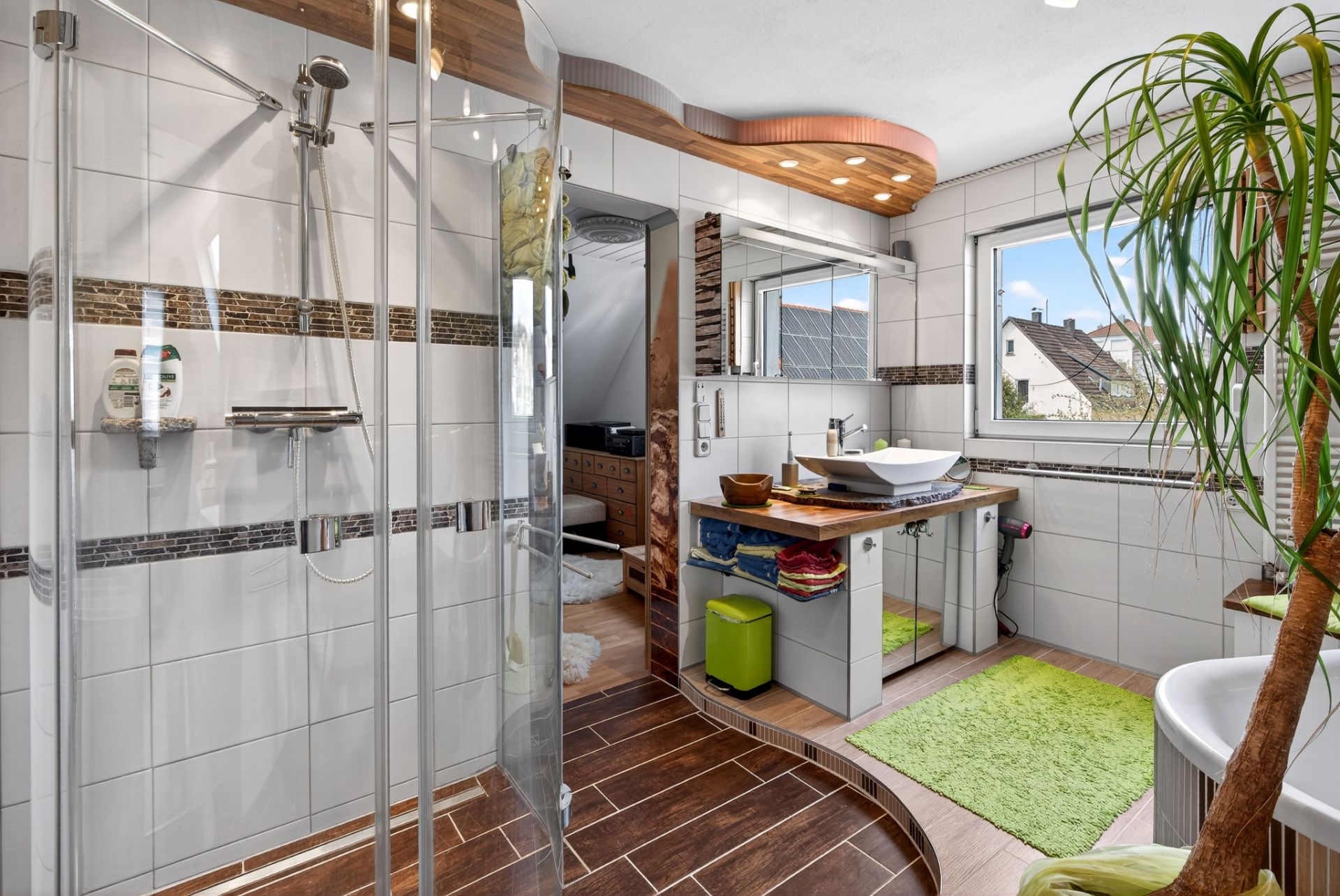 Modernes Badezimmer mit Dusche, großem Spiegel, Fenster, Pflanze und grünem Teppich.
