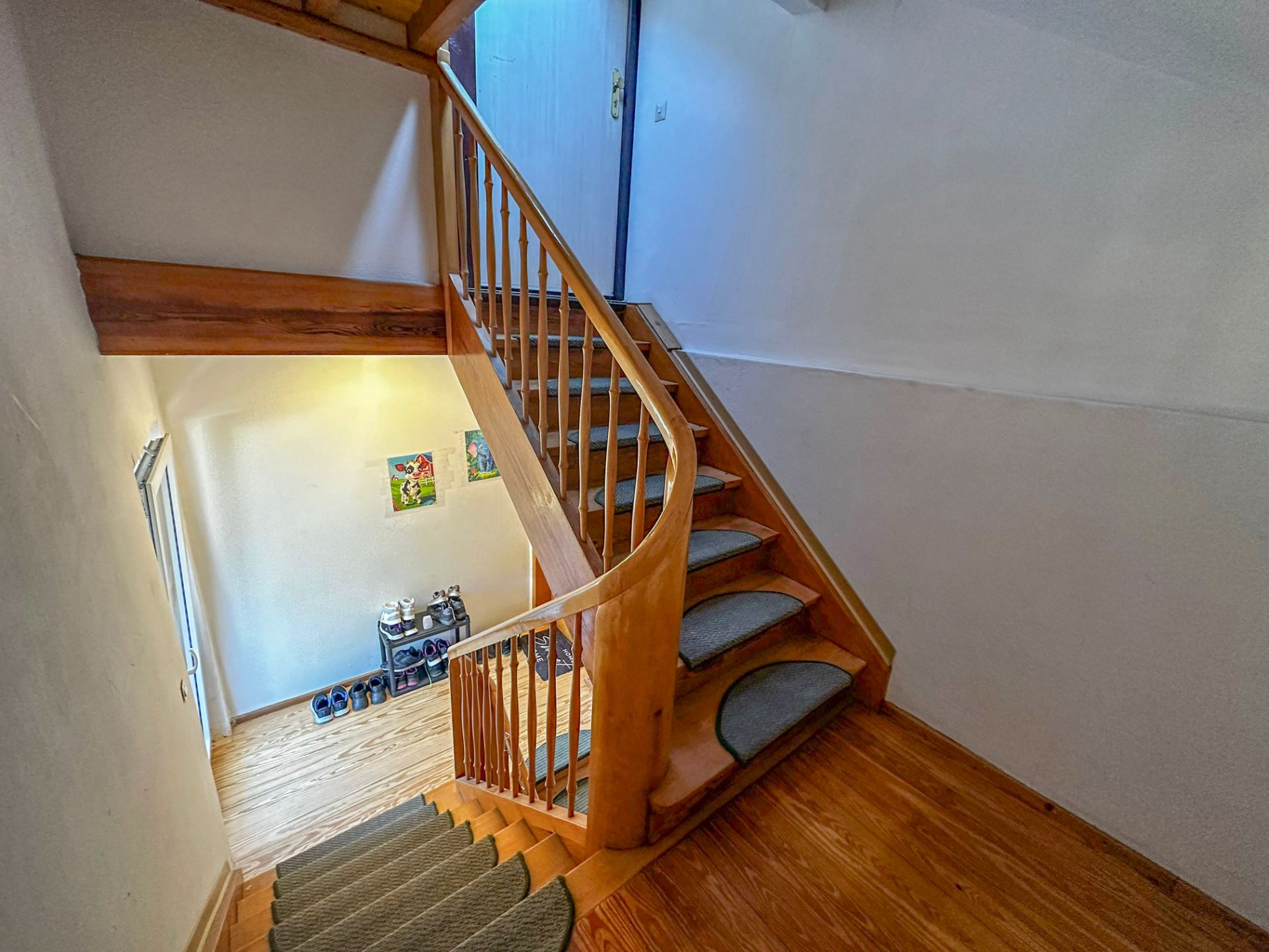 Holztreppe mit Teppichstufen in einem Hausflur, unten ein Schuhregal und Wandbilder.