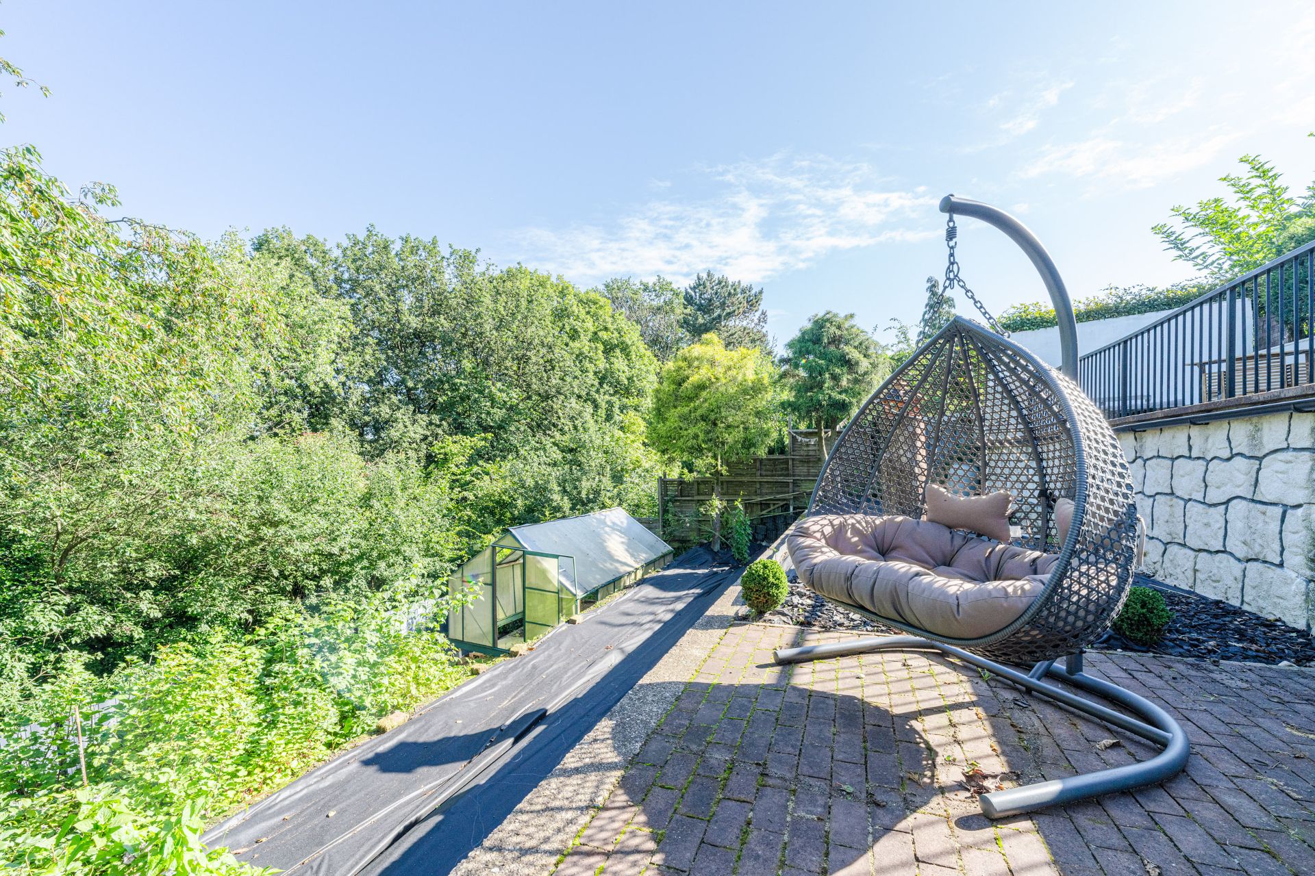 Terrasse mit Hängesessel, Blick auf Garten mit Gewächshaus und vielen Bäumen.