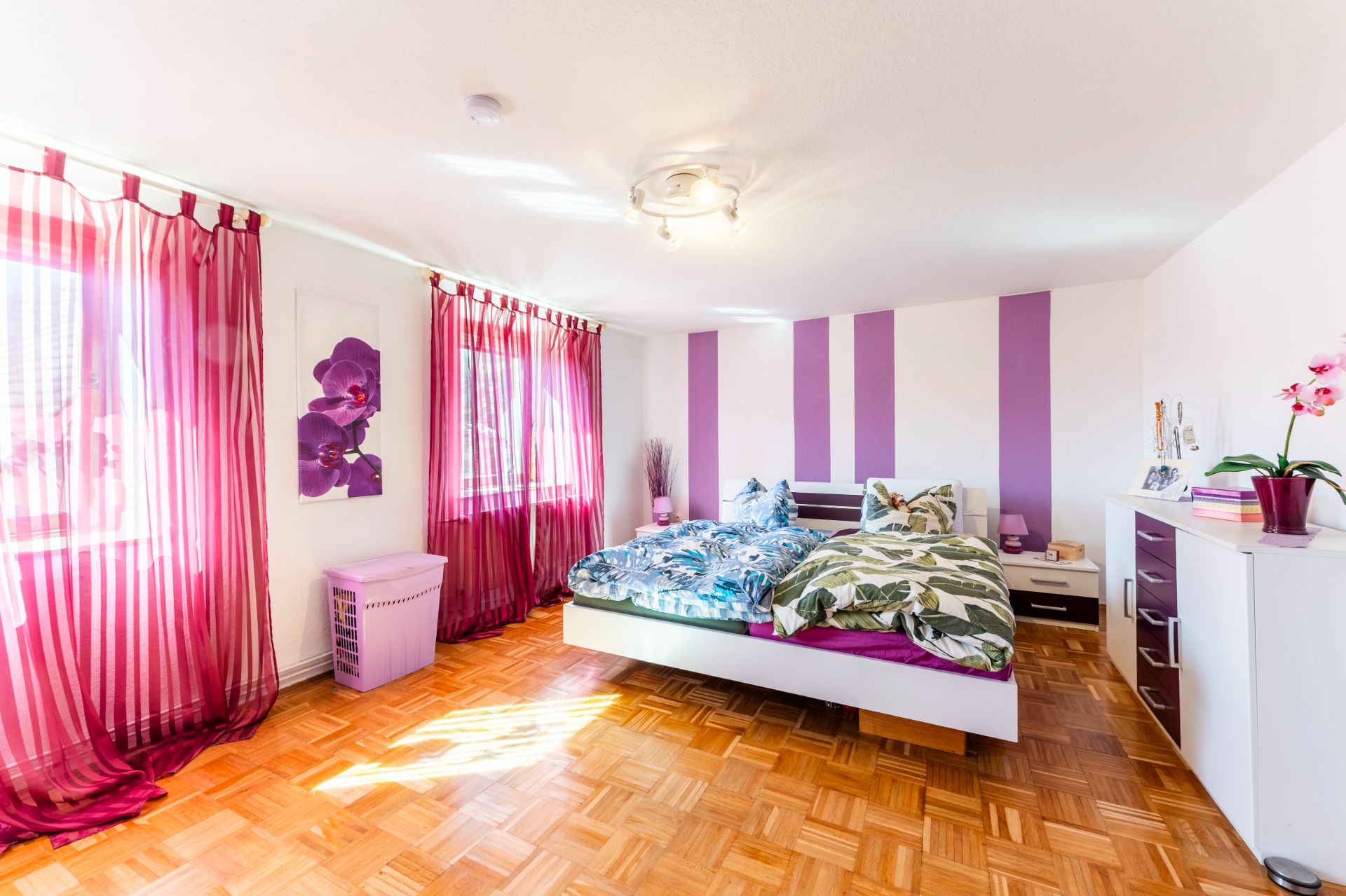 Helles Schlafzimmer mit Doppelbett, lila Vorhängen und Holzboden.
