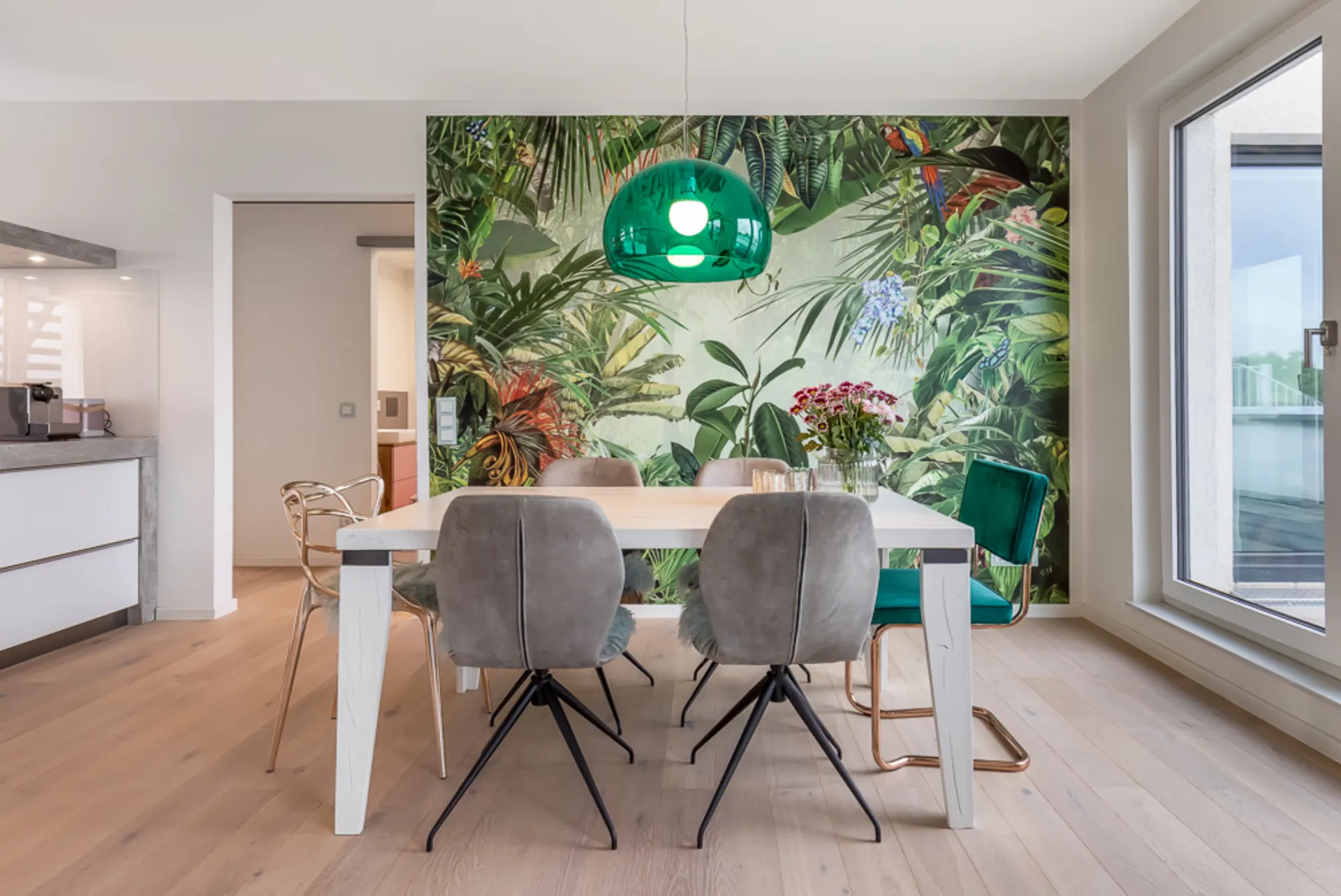 Essbereich mit bunten Stühlen, weißem Tisch und grüner Pflanzen-Tapete an der Wand.