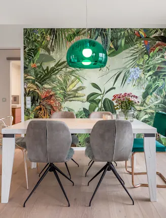 Essbereich mit bunten Stühlen, weißem Tisch und grüner Pflanzen-Tapete an der Wand.