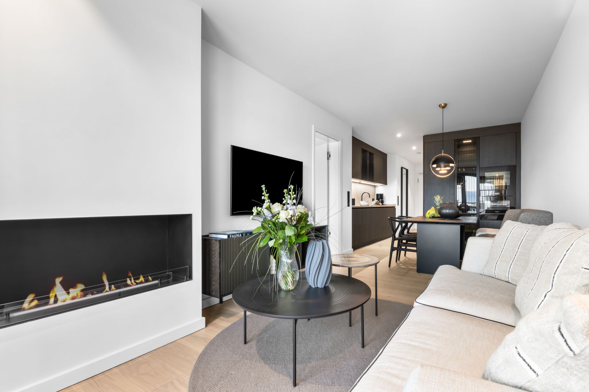 Modernes Wohnzimmer mit Sofa, Kamin, Fernseher und Esstisch, offene Küche im Hintergrund.
