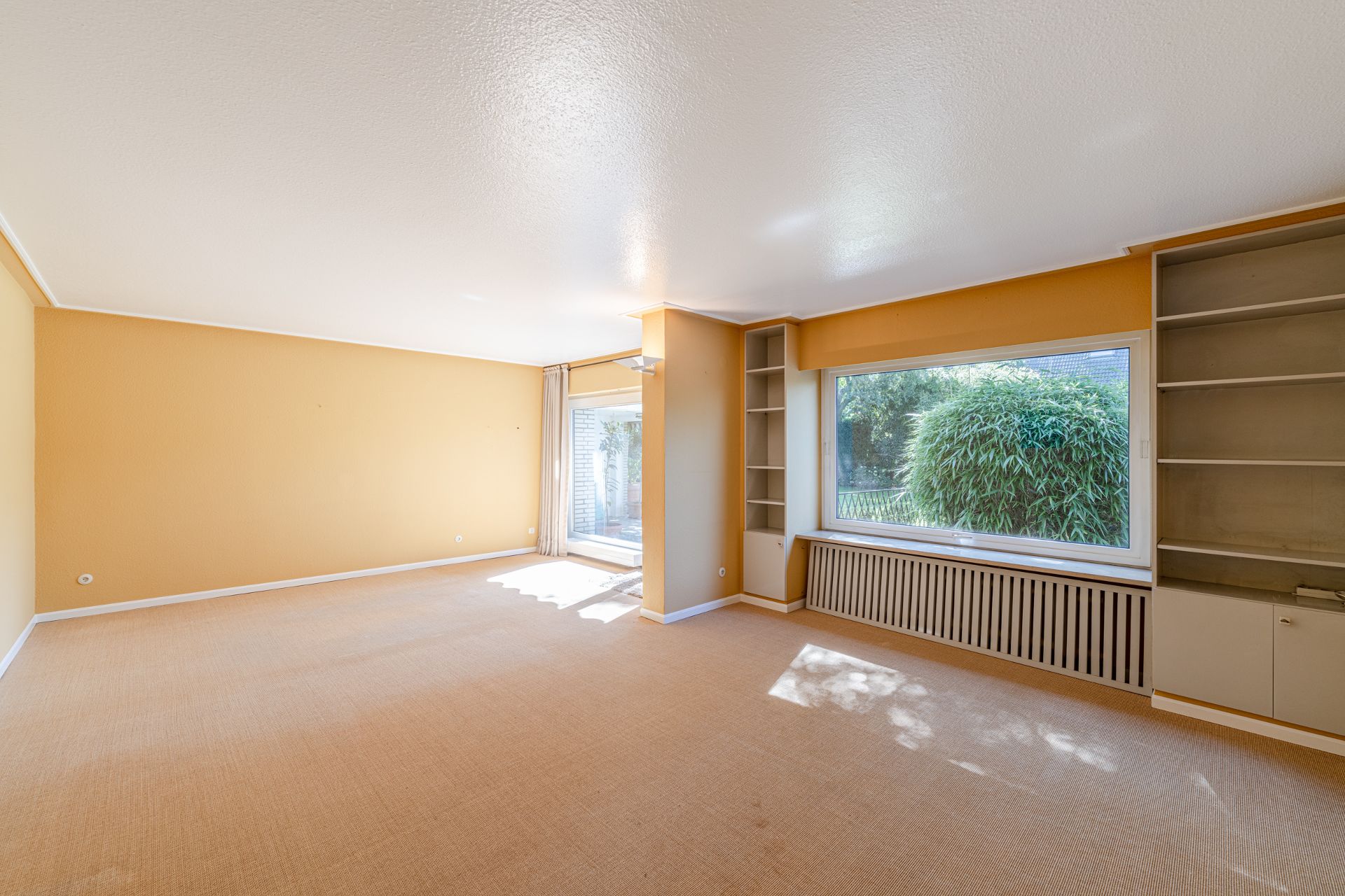 Helles, leeres Wohnzimmer mit großen Fenstern, gelben Wänden und eingebauten Regalen.