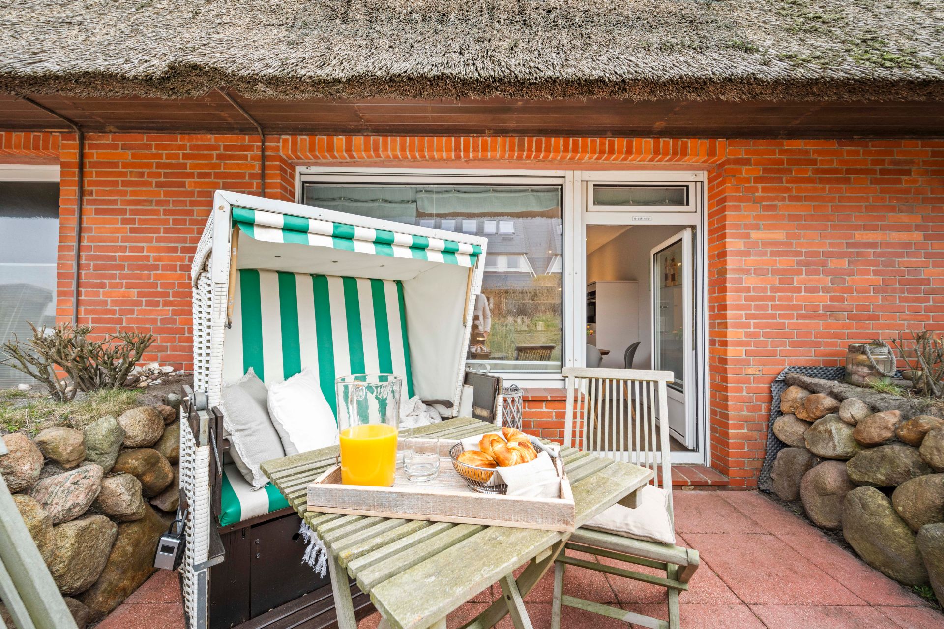 Terrasse mit Strandkorb, Holztisch, Saft und Croissants vor Backsteinwand.
