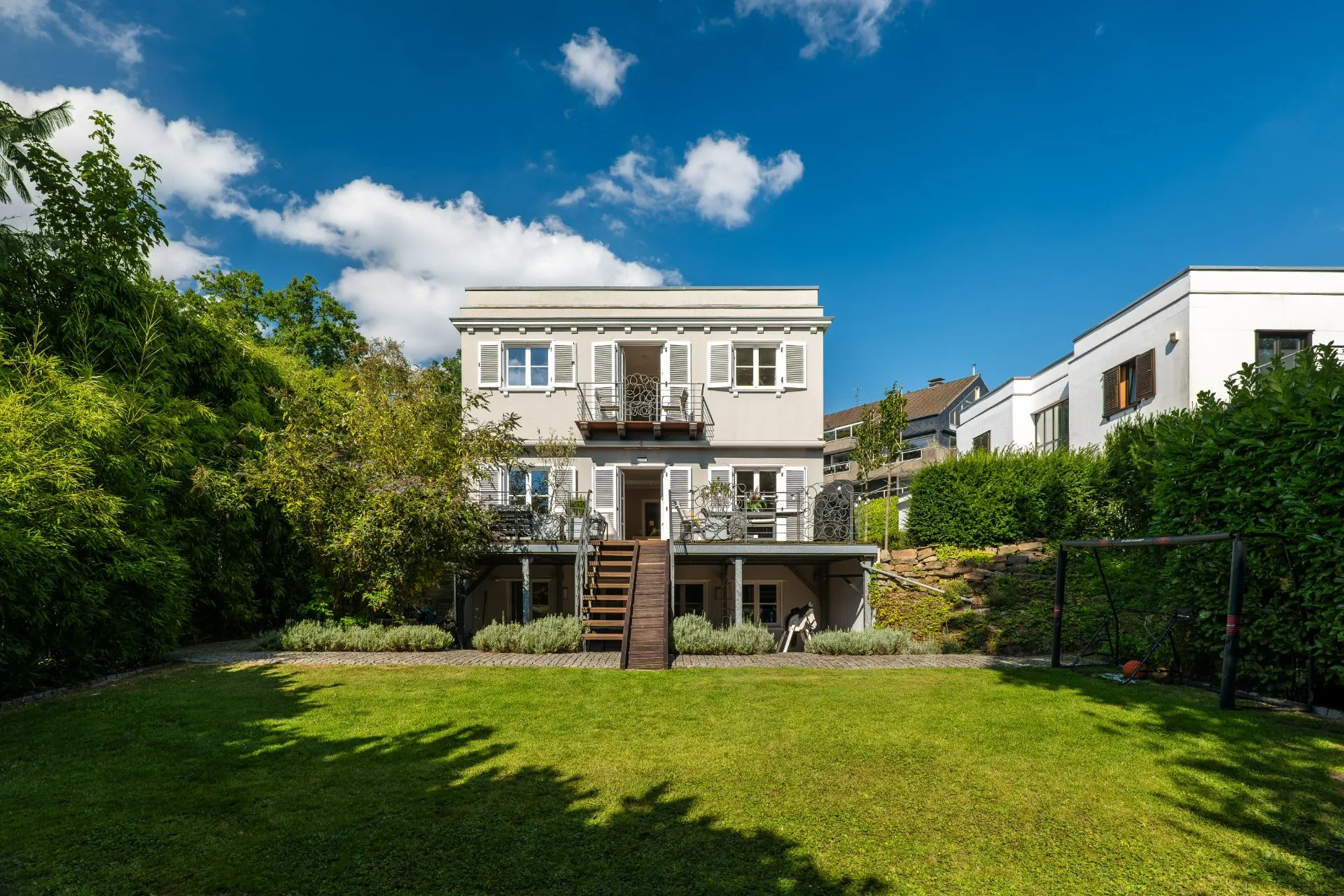 Zweistöckiges Wohnhaus mit Balkon und Garten, umgeben von Bäumen und Rasen.