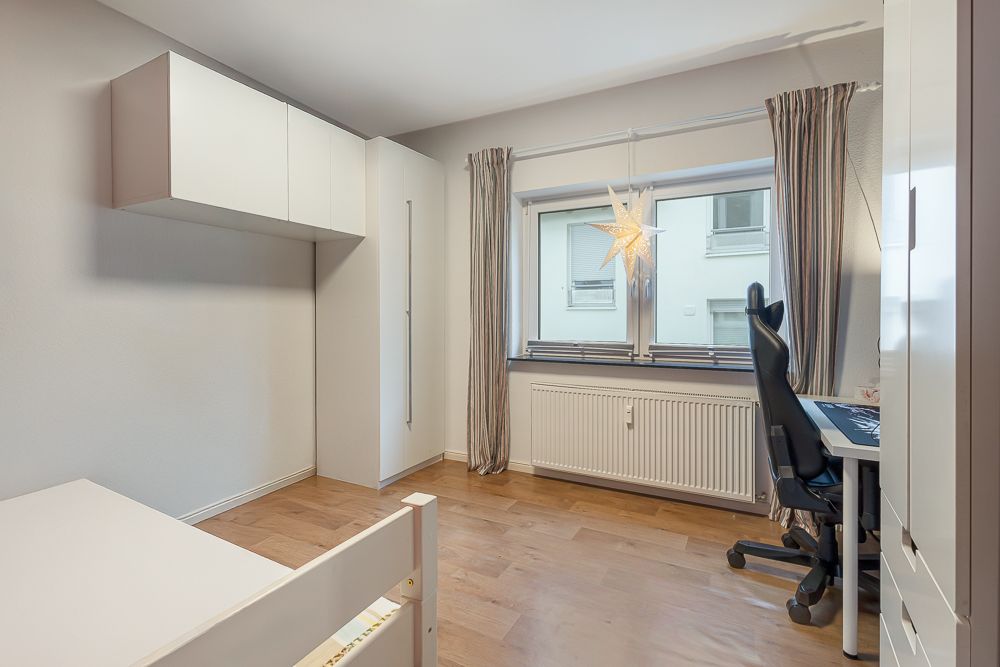 Helles Zimmer mit Schreibtisch, Stuhl, Bett, Schränken und Fenster mit Vorhängen.