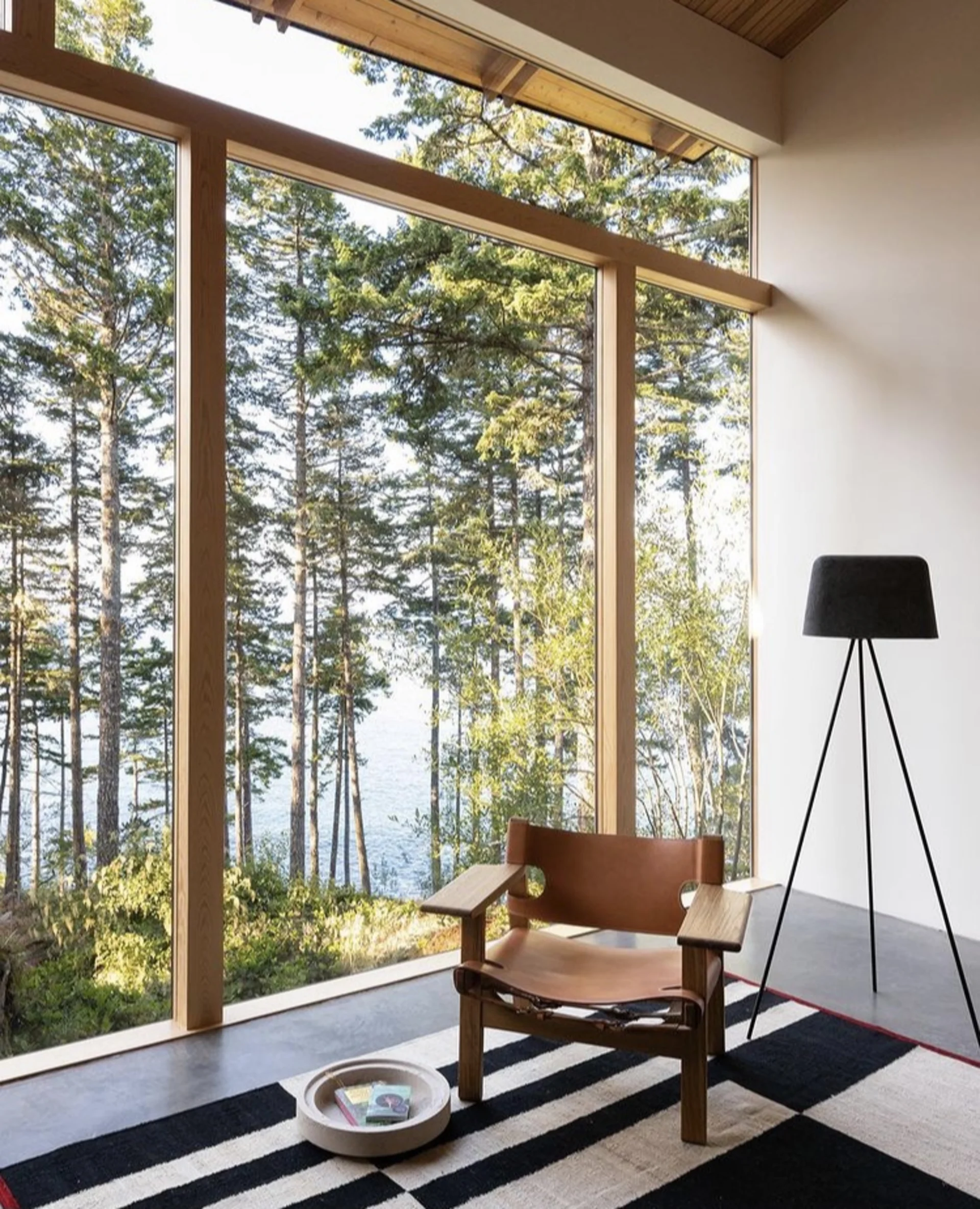 Holzstuhl und Stehlampe vor großer Fensterfront mit Blick in einen Wald.