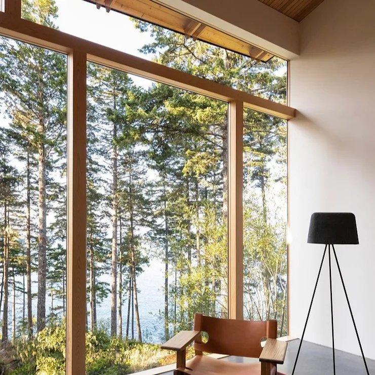 Holzstuhl und Stehlampe vor großer Fensterfront mit Blick in einen Wald.