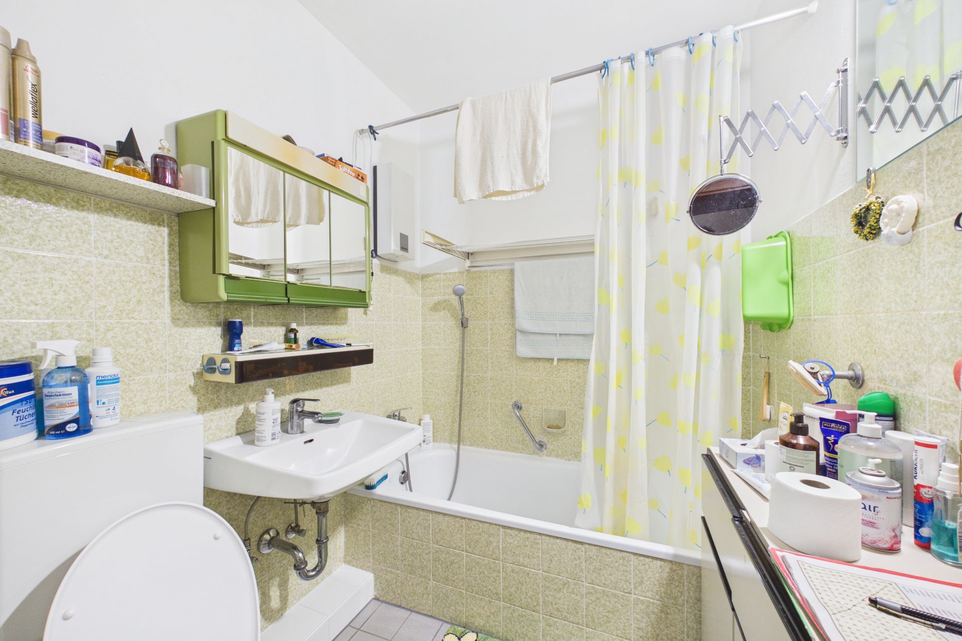 Kleines Badezimmer mit Badewanne, Duschvorhang, Waschbecken, Toilette und vielen Pflegeprodukten.