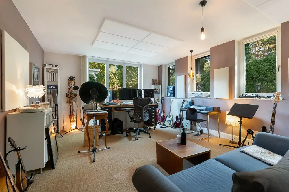 Wohnzimmer mit Schreibtisch, Computern, Musikinstrumenten und Sofa, große Fenster mit Blick ins Grüne.