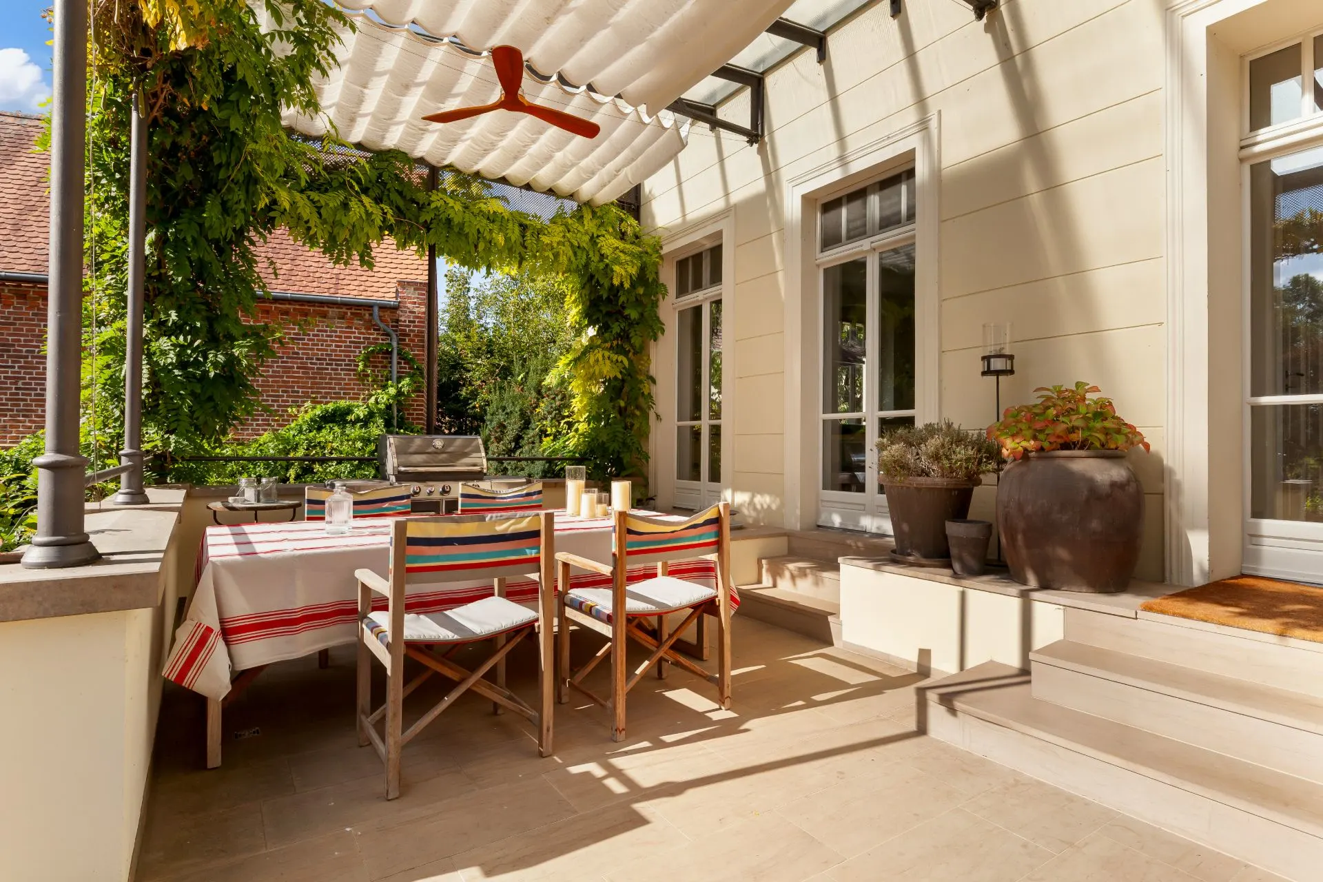Sonnige Terrasse mit Esstisch, Stühlen, Markise und Pflanzen, angrenzend an ein Wohnhaus.