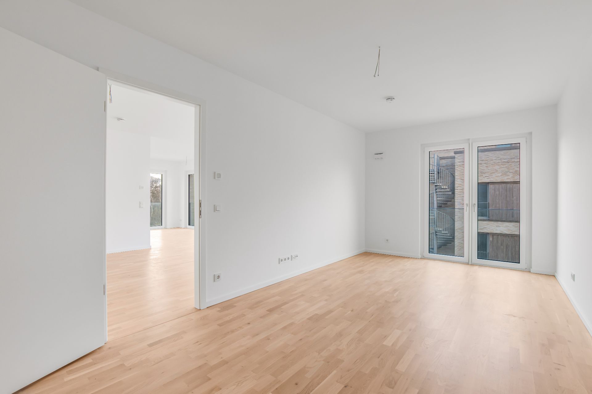 Heller leerer Raum mit Holzfußboden, weißen Wänden und großen Fenstertüren zum Balkon.
