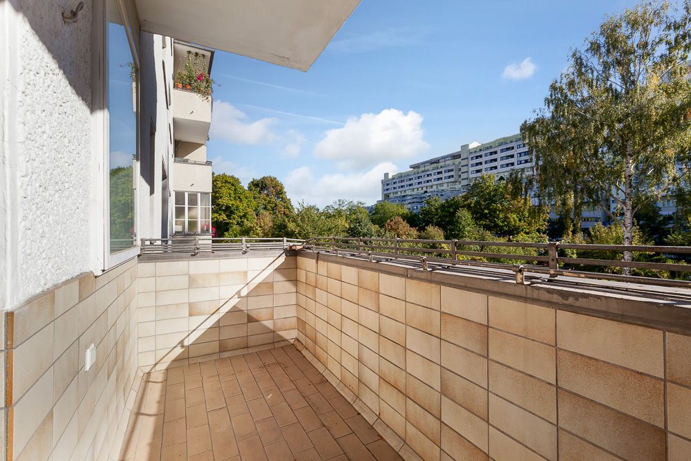 Balkon mit Fliesenboden und Ausblick auf Bäume und ein großes Wohngebäude im Hintergrund.