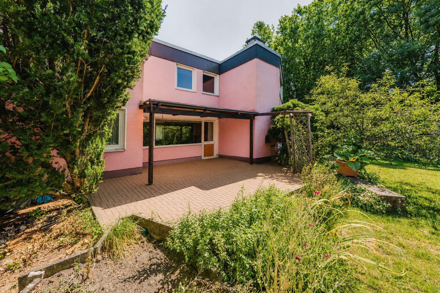 Zweistöckiges rosa Haus mit großer Terrasse und Garten, umgeben von Bäumen und Sträuchern.