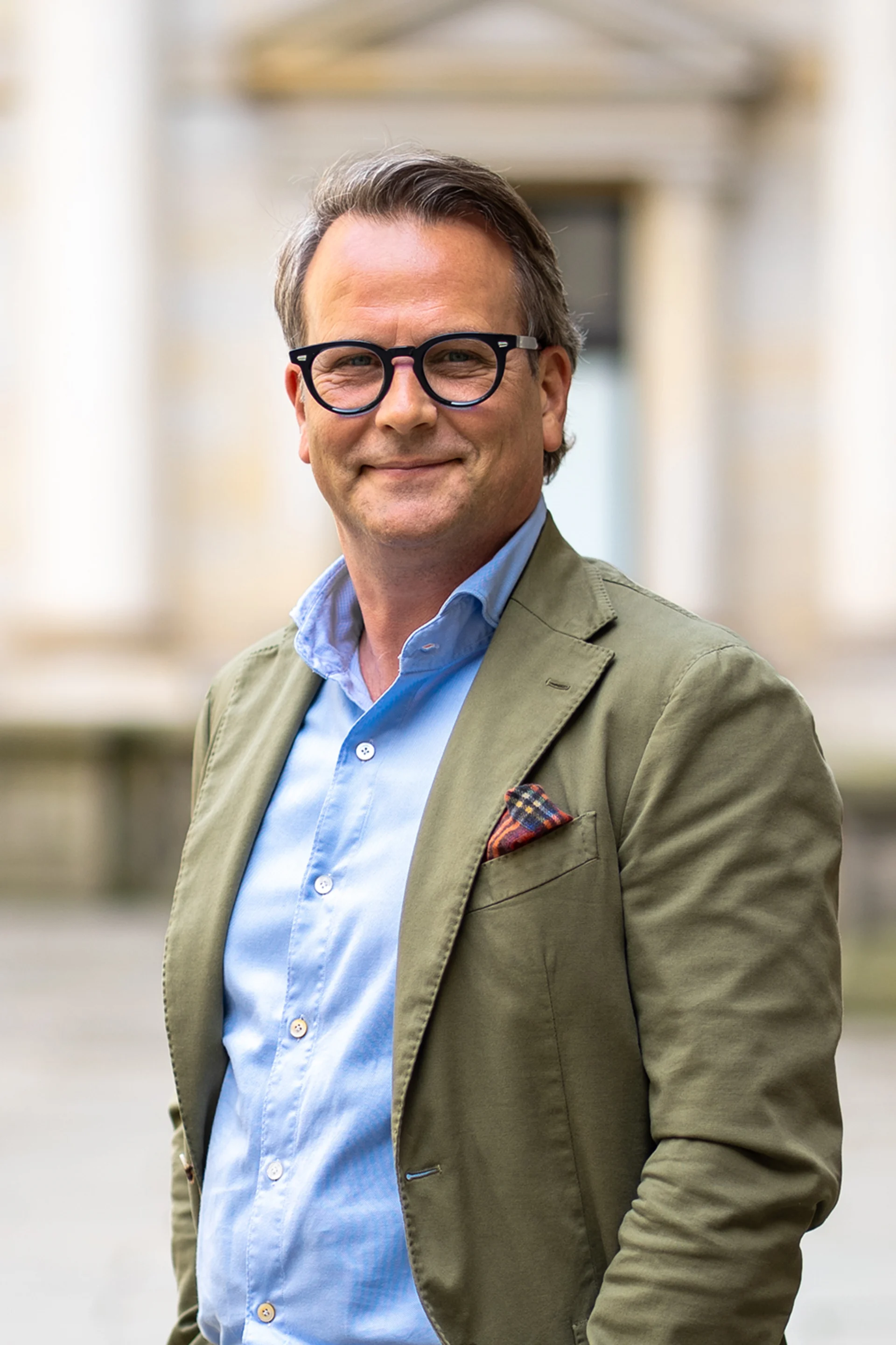 Ein Mann mit Brille im grünen Sakko steht draußen und lächelt in die Kamera.